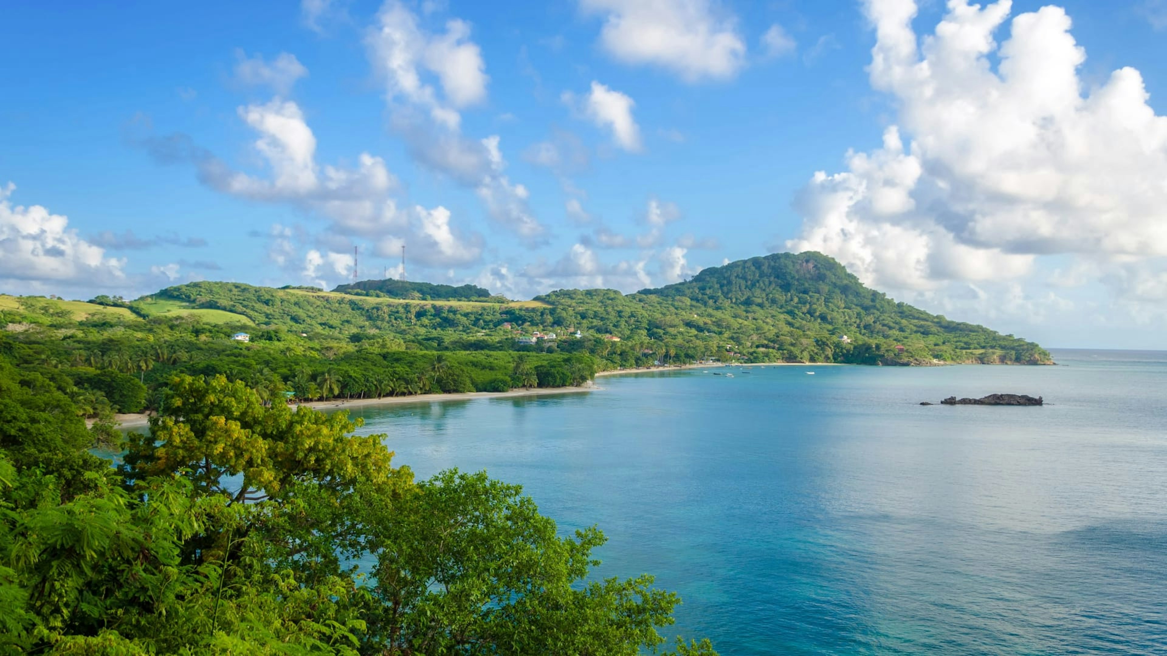 A lush green coastline with hills, trees, and a sandy beach borders a calm blue ocean under a partly cloudy sky.