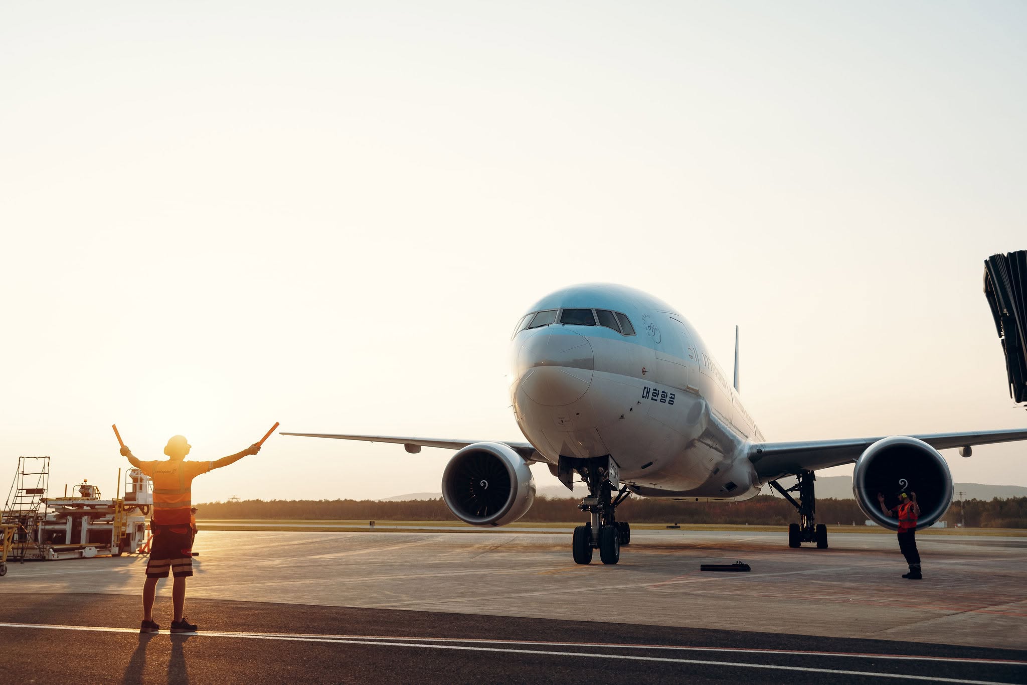 Un membre du personnel au sol guide un gros avion commercial qui roule sur la piste au coucher du soleil ; on aperçoit à proximité un autre employé et du matériel aéroportuaire.