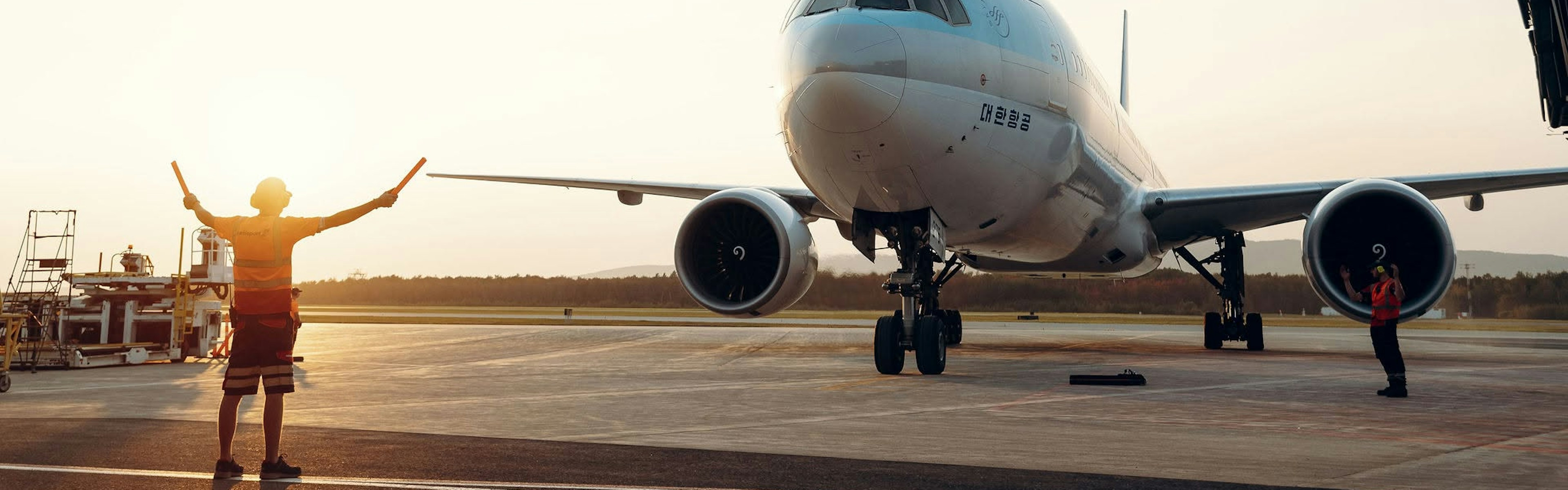 Un membre du personnel au sol guide un gros avion commercial qui roule sur la piste au coucher du soleil ; on aperçoit à proximité un autre employé et du matériel aéroportuaire.