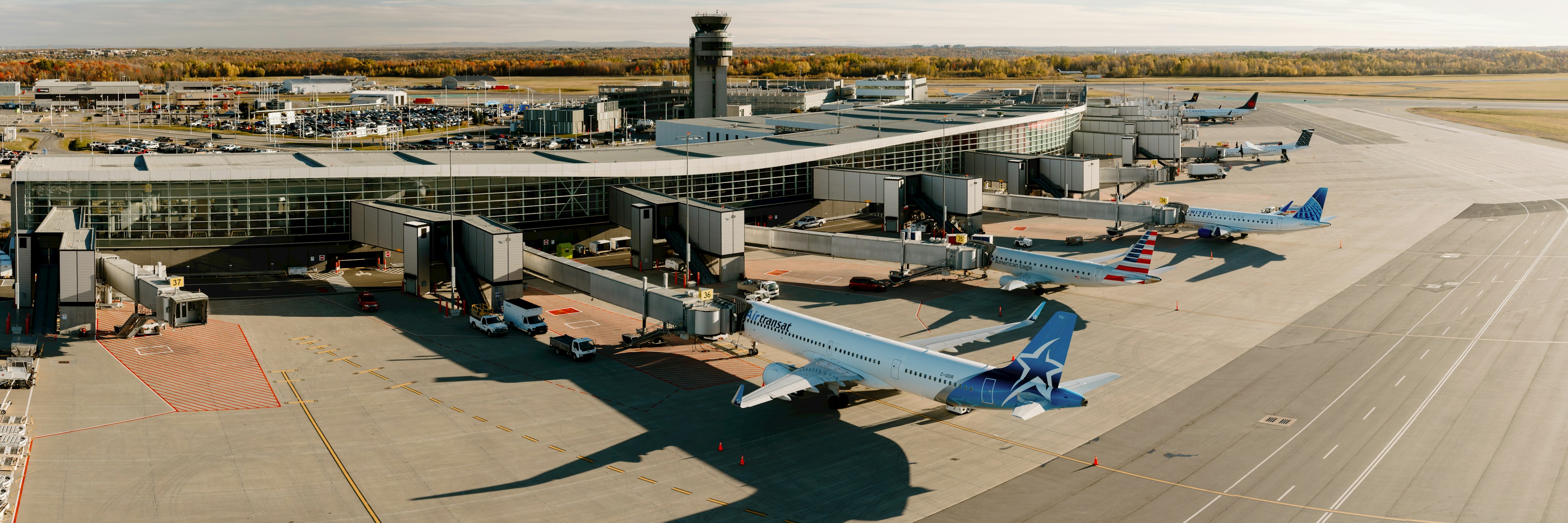 Vue aérienne d'un terminal d'aéroport avec plusieurs avions stationnés aux portes d'embarquement, des passerelles d'embarquement raccordées, et une tour de contrôle en arrière-plan, sous un ciel dégagé.
