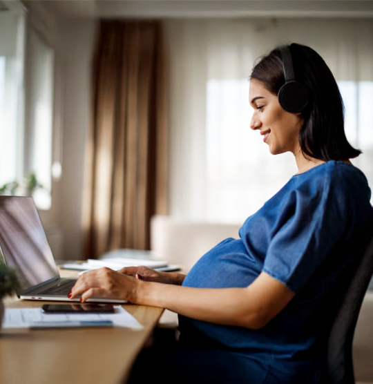 Schwangerschaft Wissen Bild Smiling pregnant woman sitting in front of a laptop and wearing headphones