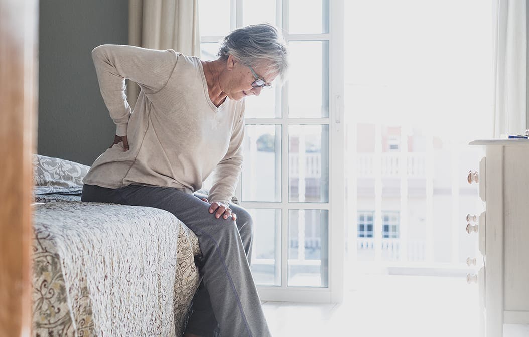 older woman sitting on a bed, holding her lower back in pain