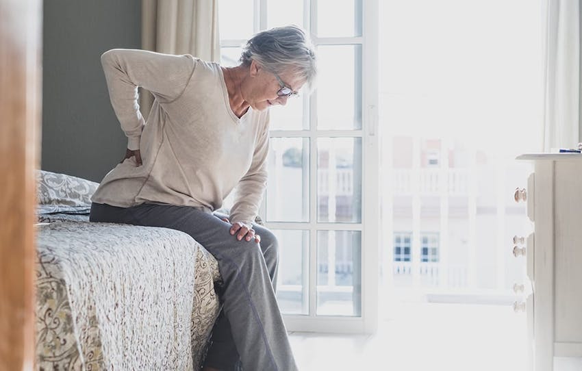 older woman sitting on a bed, holding her lower back in pain
