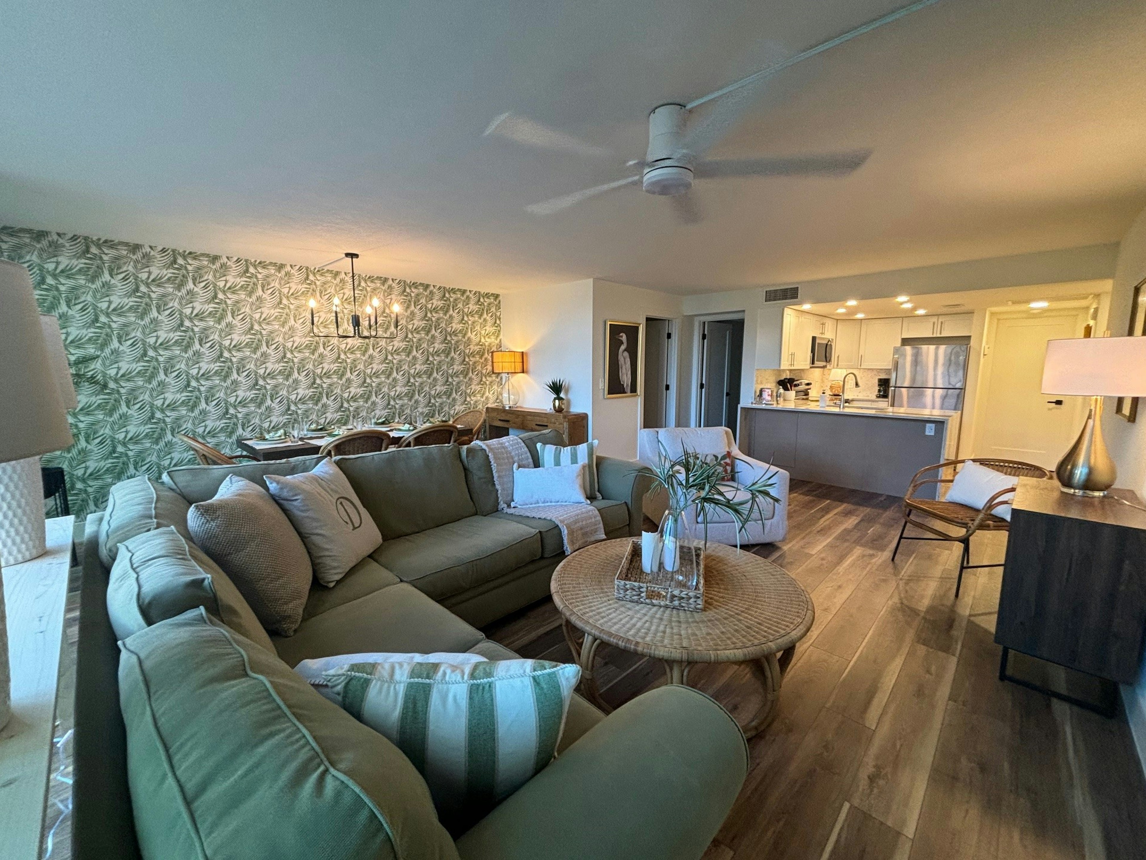 Relax on your plush sectional sofa in this open-concept living space featuring tropical-inspired decor and a sleek kitchen with modern appliances.