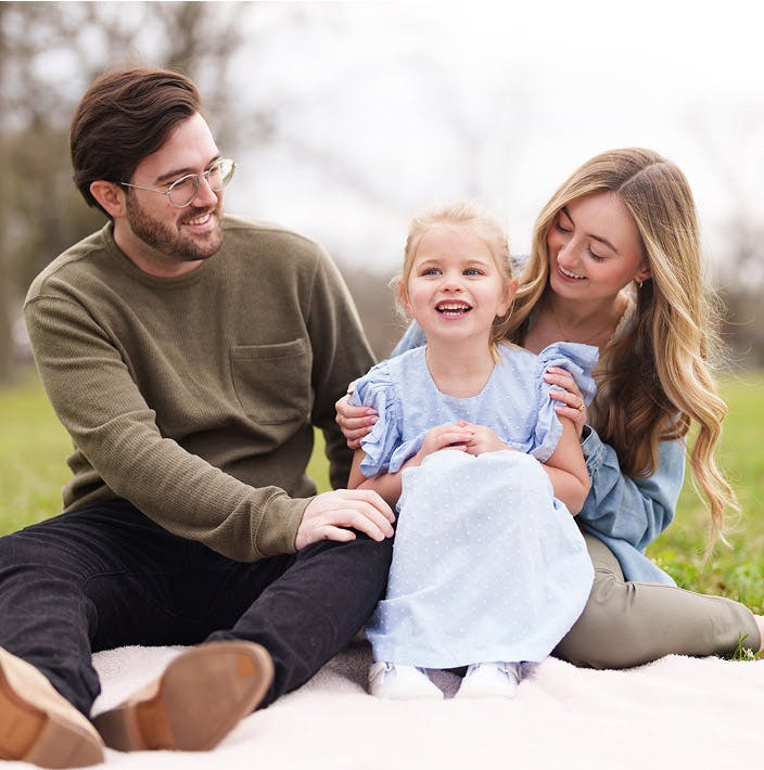 man, woman, and little girl sitting on a blanket in a park