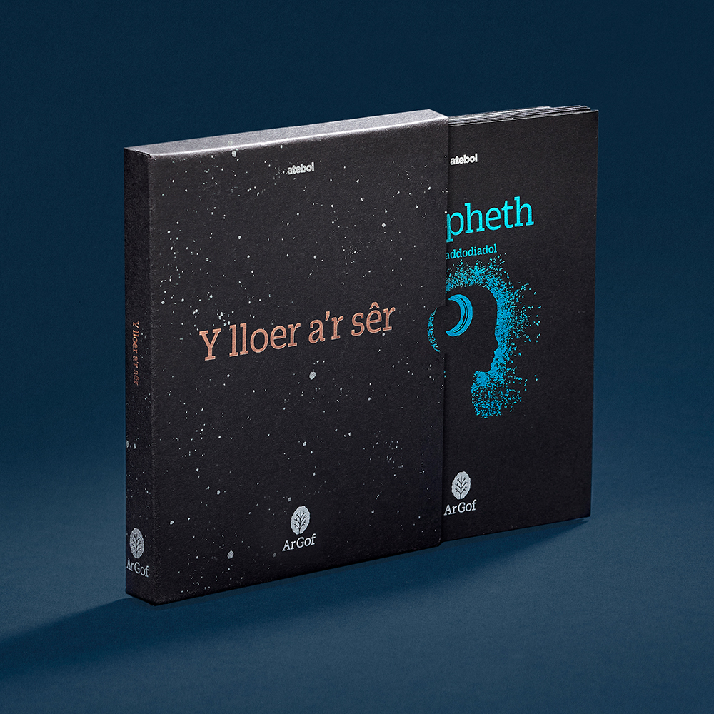 Dark grey book with luminous blue illustration half in gold foiled dark grey sleeve, shot on dark blue background