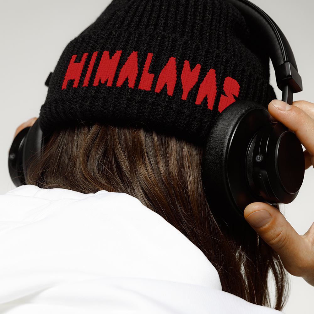 Back of young man's head wearing black beanie with red embroidered Himalayas logo and headphones while listening to music
