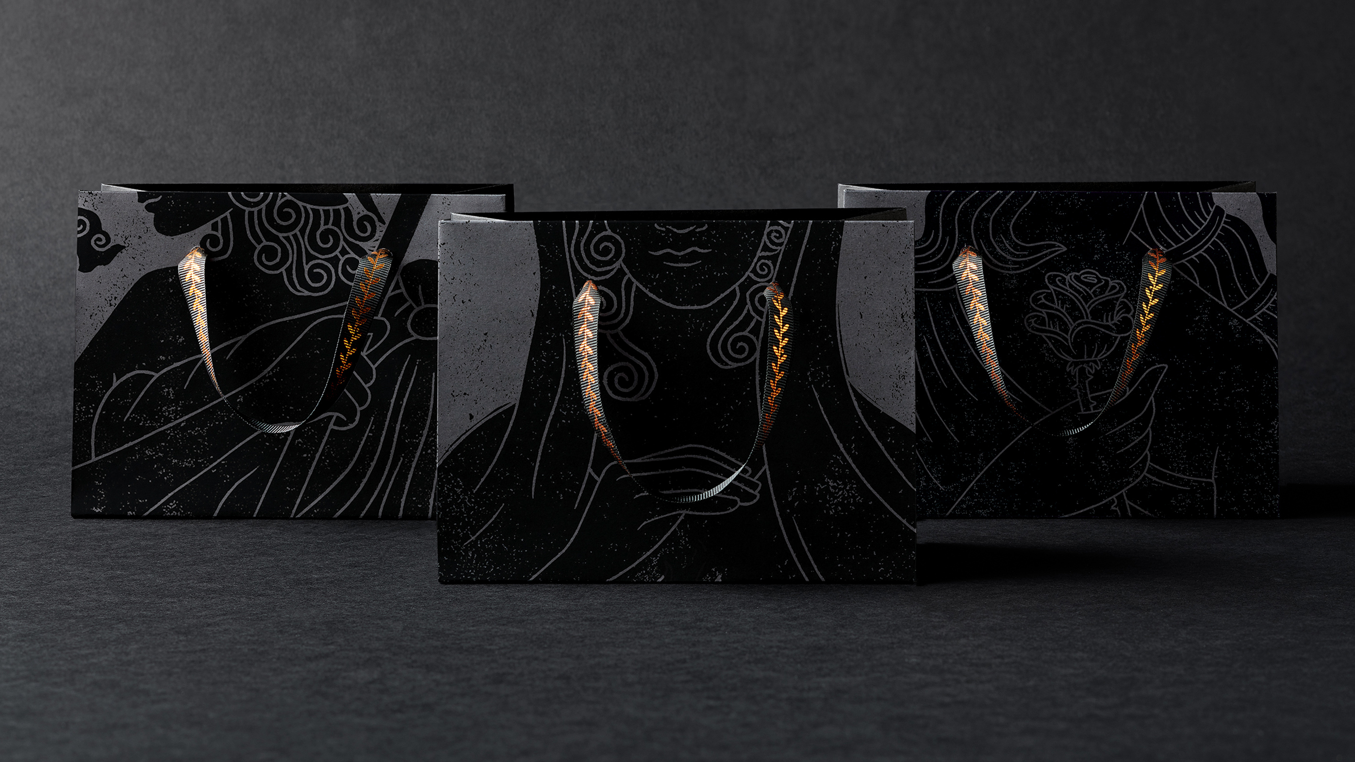 Three luxury paper bags on dark grey backdrop with black and grey Greek goddess illustrations and black ribbon handles with bronze olive branch pattern