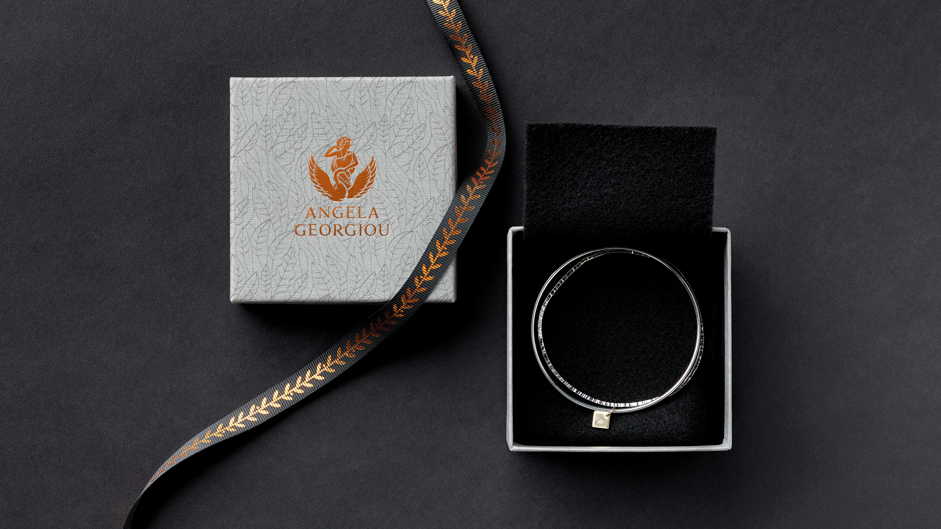 Opened jewellery box with silver bangles, bronze Angela Georgiou logo foiled on leaf-patterned lid, and black ribbon with bronze olive branch pattern