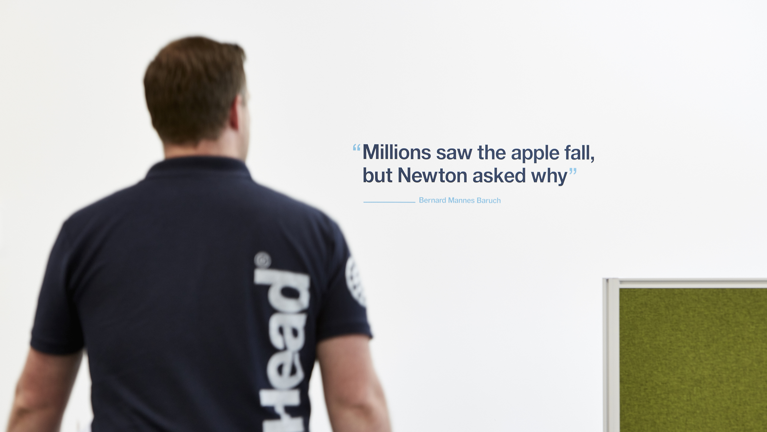 Man in bigHead t-shirt walking past a white wall in office with blue and cyan vinyl quote reading "millions saw the apple fall, but Newton asked why"