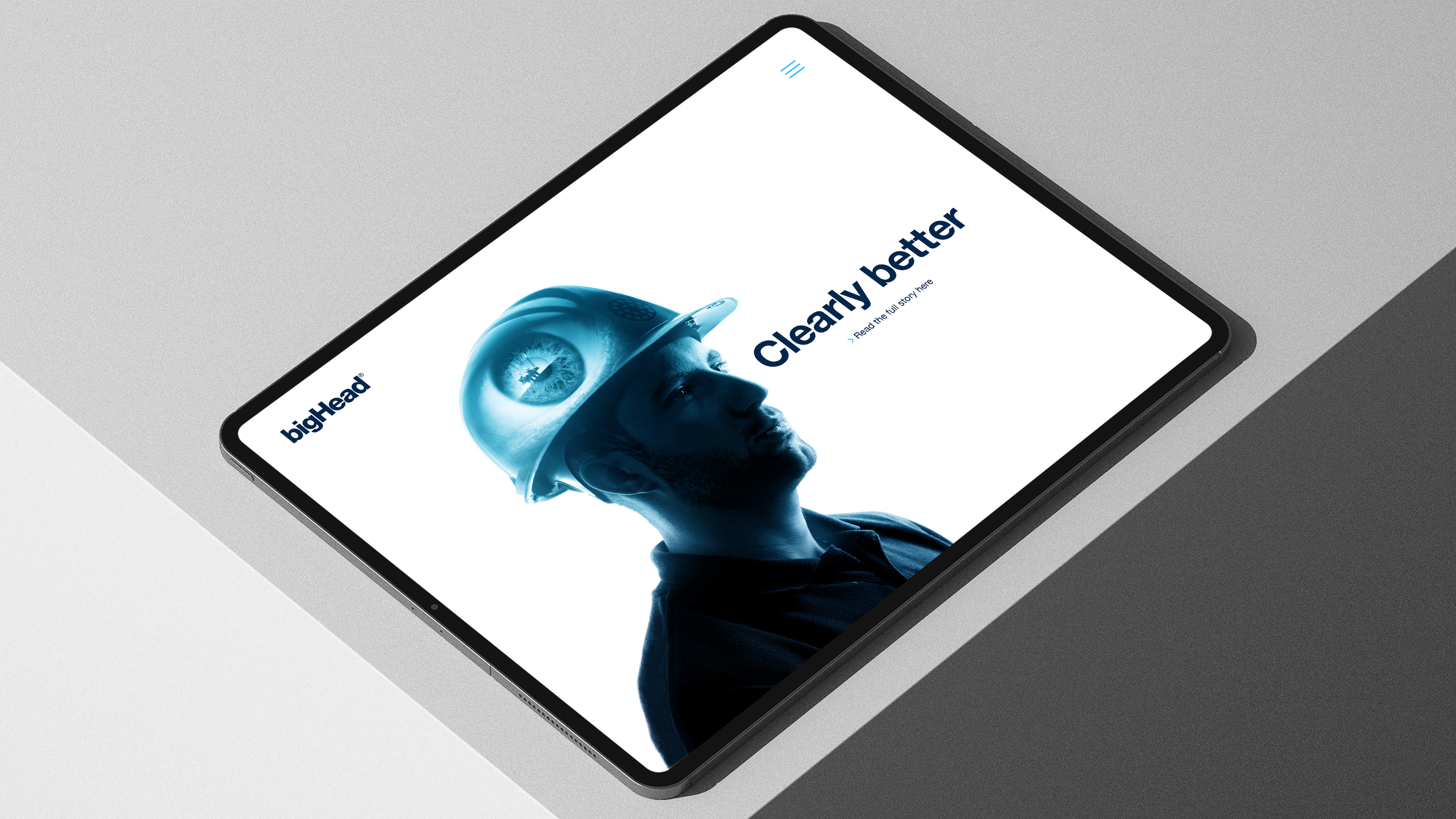 iPad on grey surface showing bigHead webpage with blue double exposure of engineer and eye alongside logo and words "clearly better" in dark blue