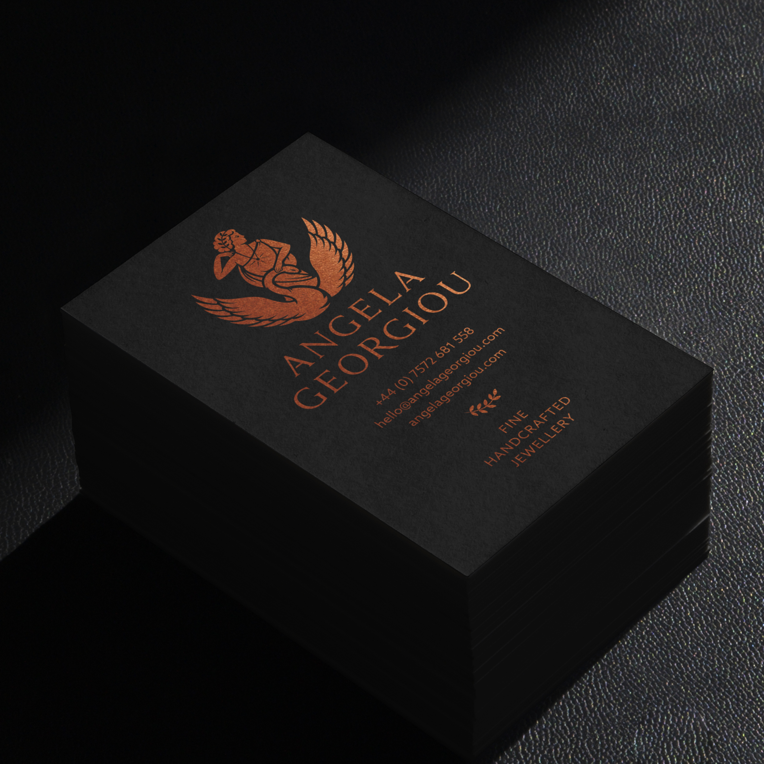 Stack of black business cards with bronze foiling of Angela Georgiou's Greek mythology inspired logo and details, on black leather surface in low lighting