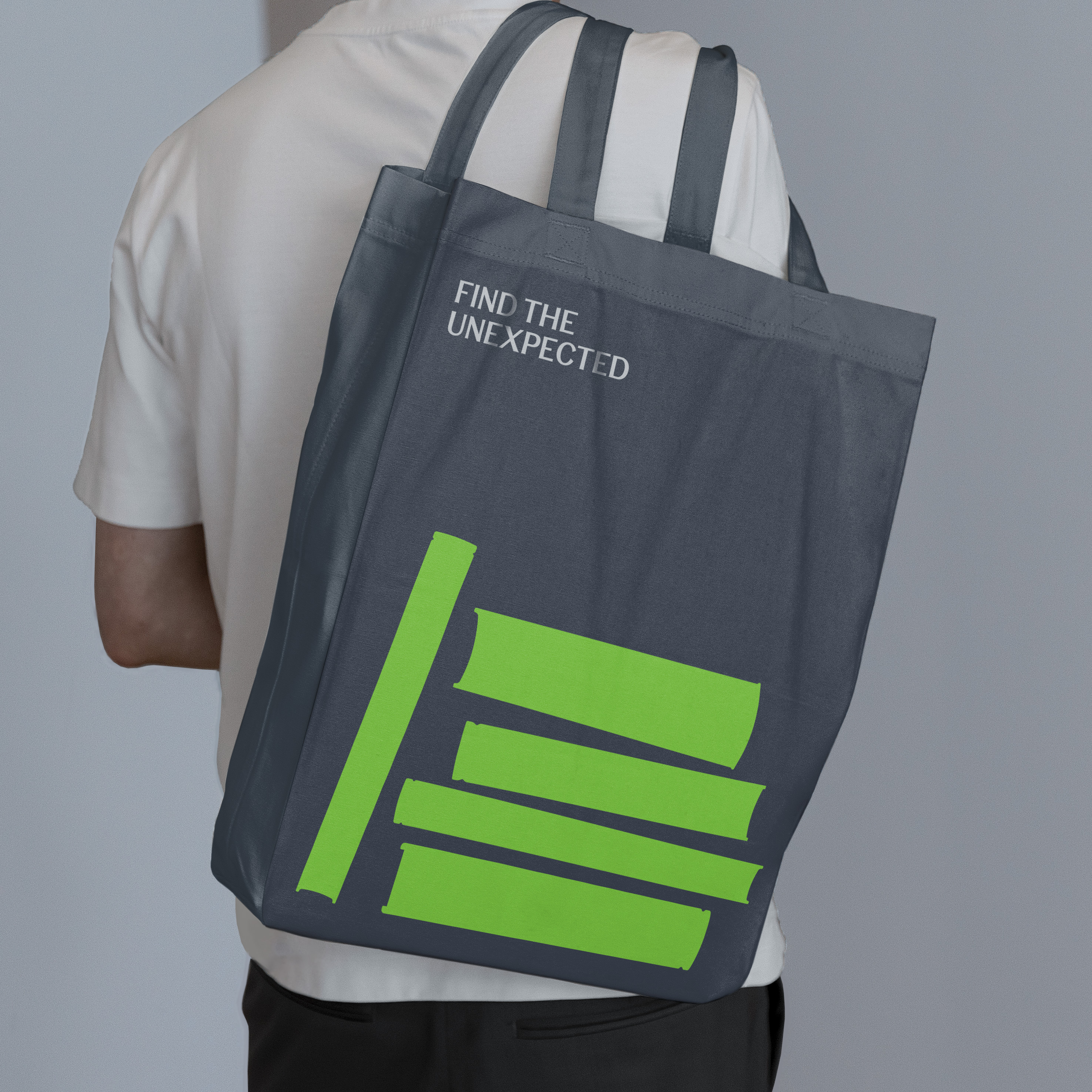 Back view of man in white T-shirt carrying dark grey tote bag with bright green book spine graphics and 'Find the unexpected' text