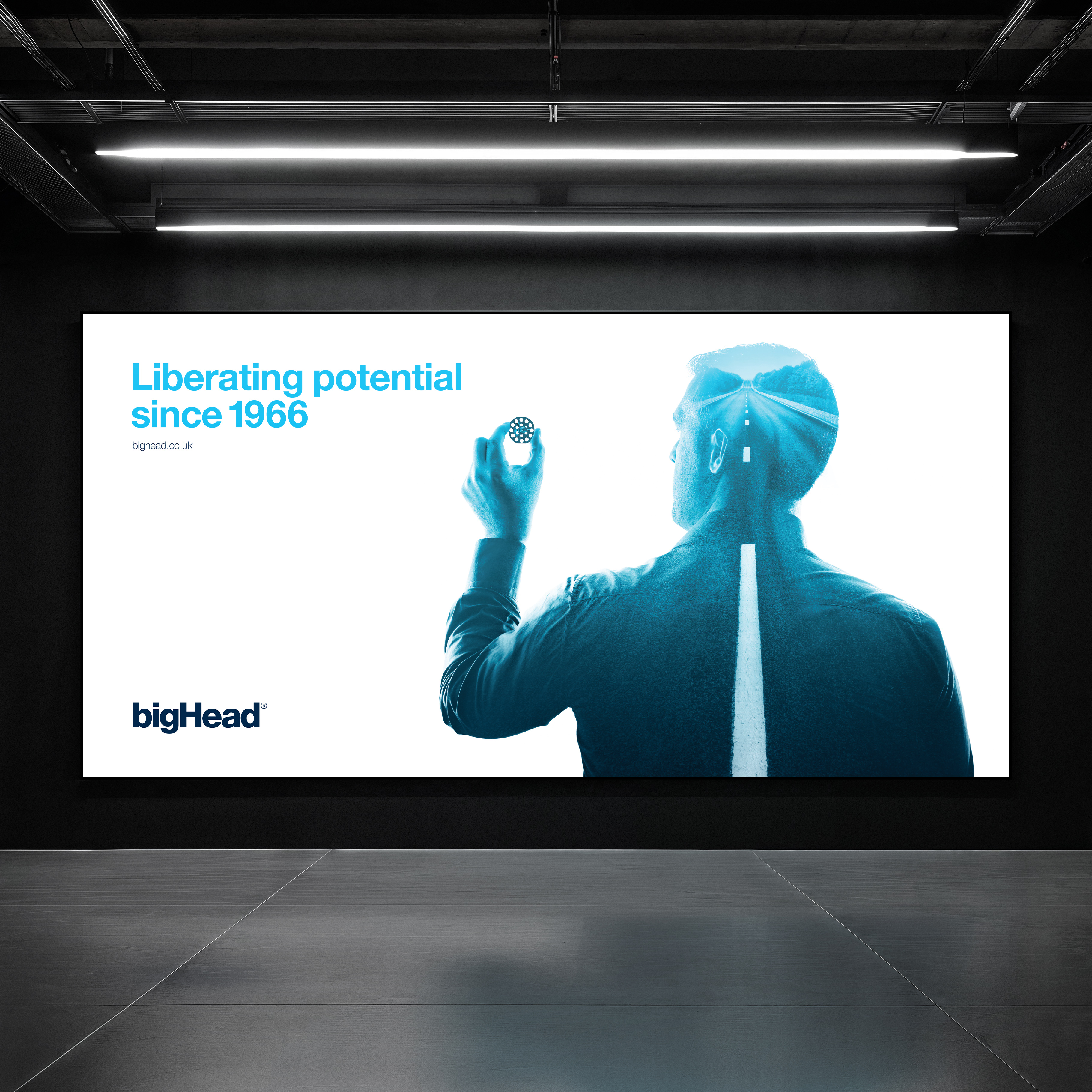 Digital billboard in industrial building showing blue double exposure of man holding bigHead with road image, logo and strapline in brand blues