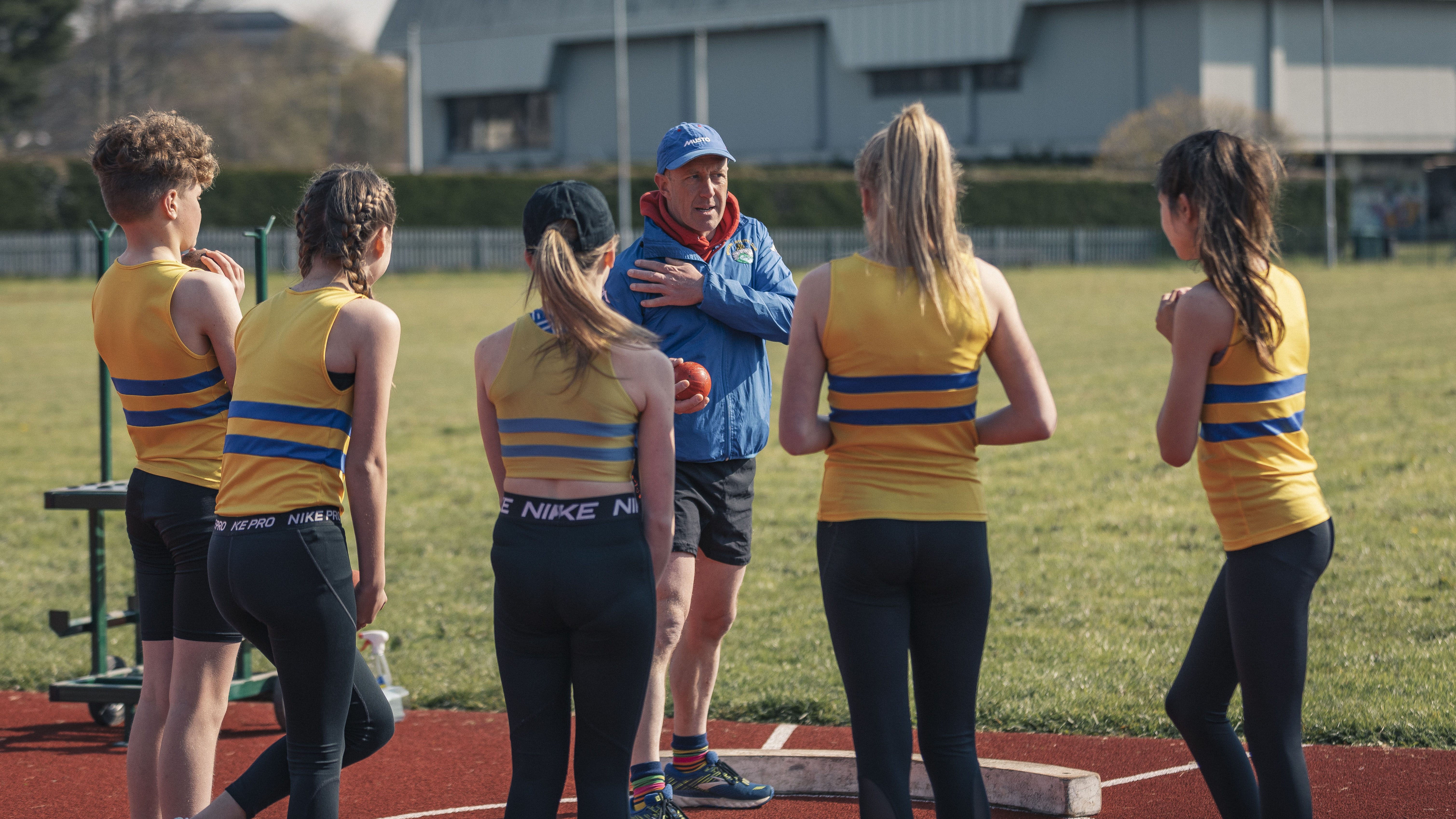 Group of children in Bridgend Athletics jerseys watching coach’s technique demonstration on sports field on sunny day