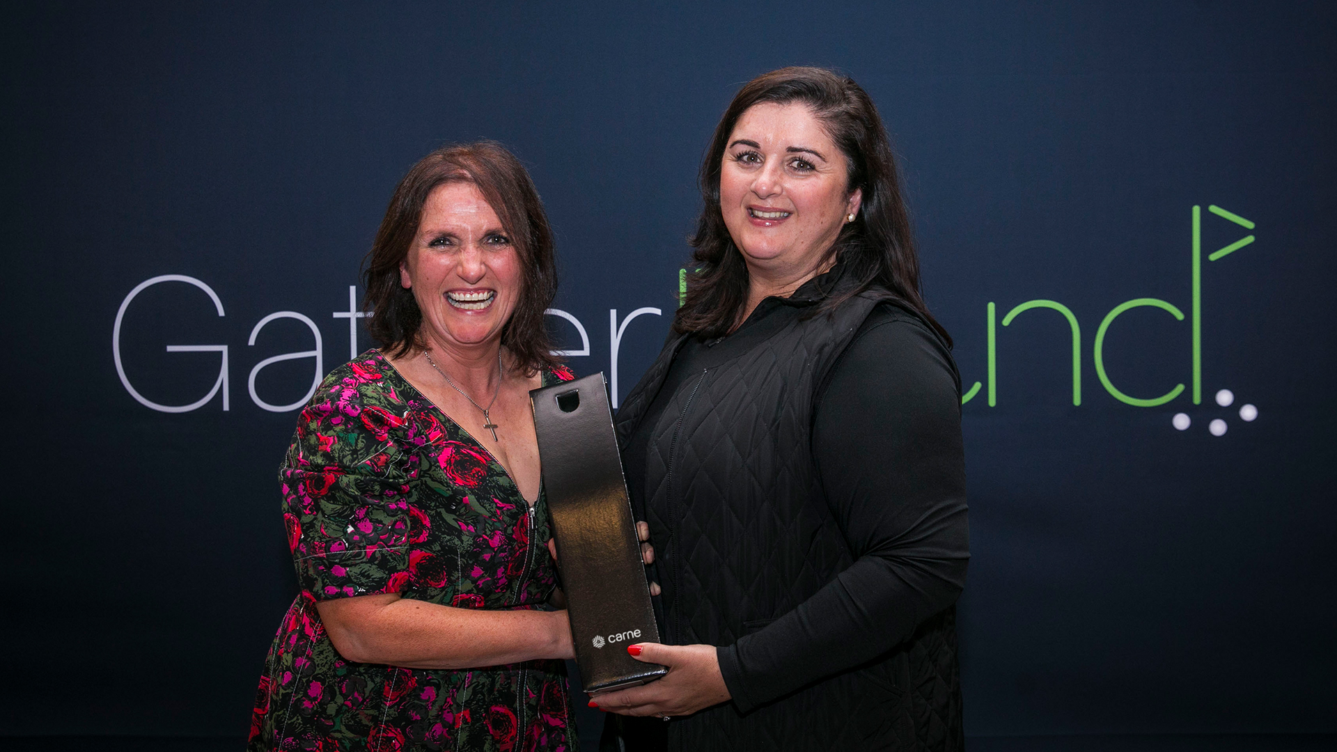 Two women celebrating prize at Carne Group GatherRound golf event with green and white logo on dark grey backdrop behind them