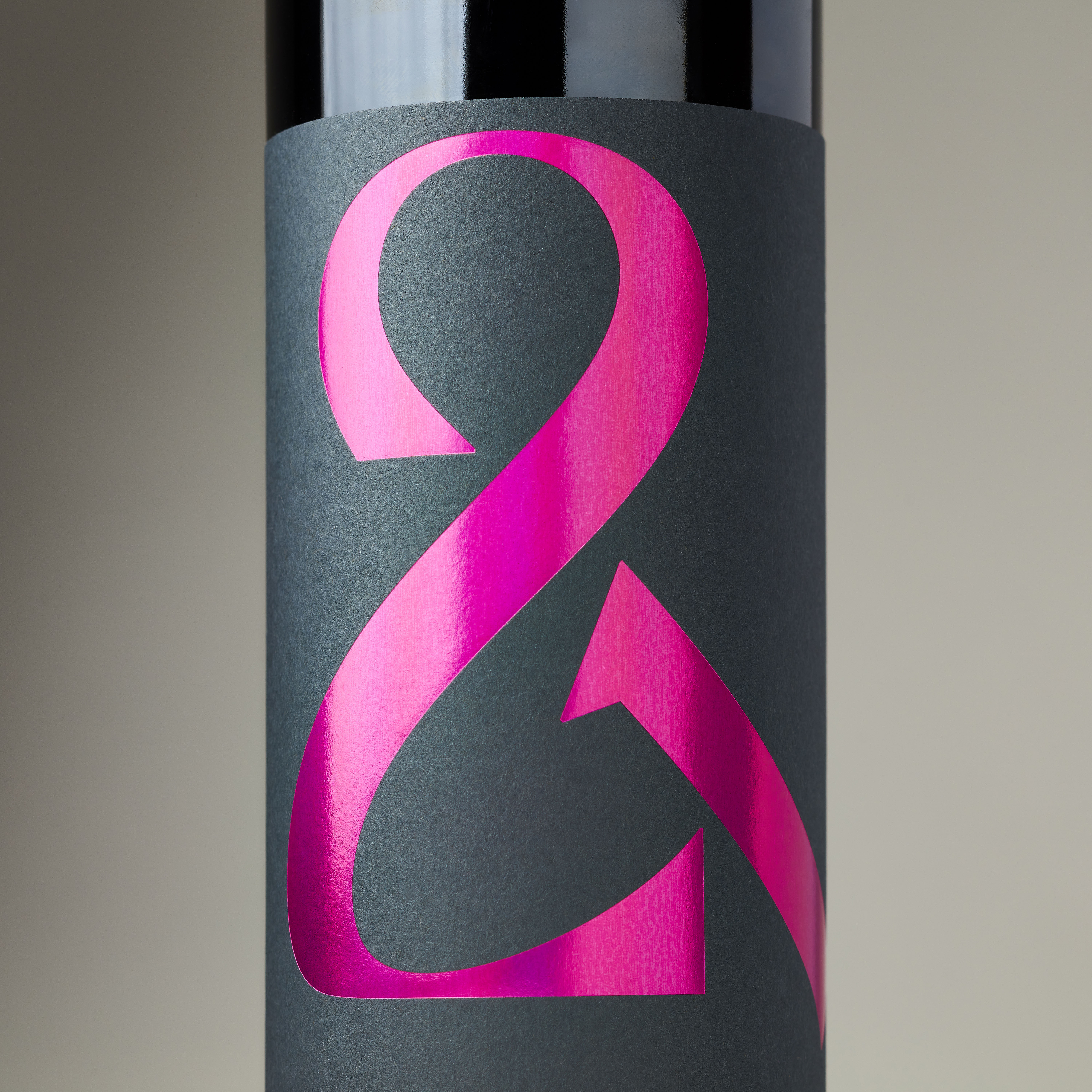 Close-up of red wine bottle label in dark grey with holographic hot pink ampersand logo under studio lighting