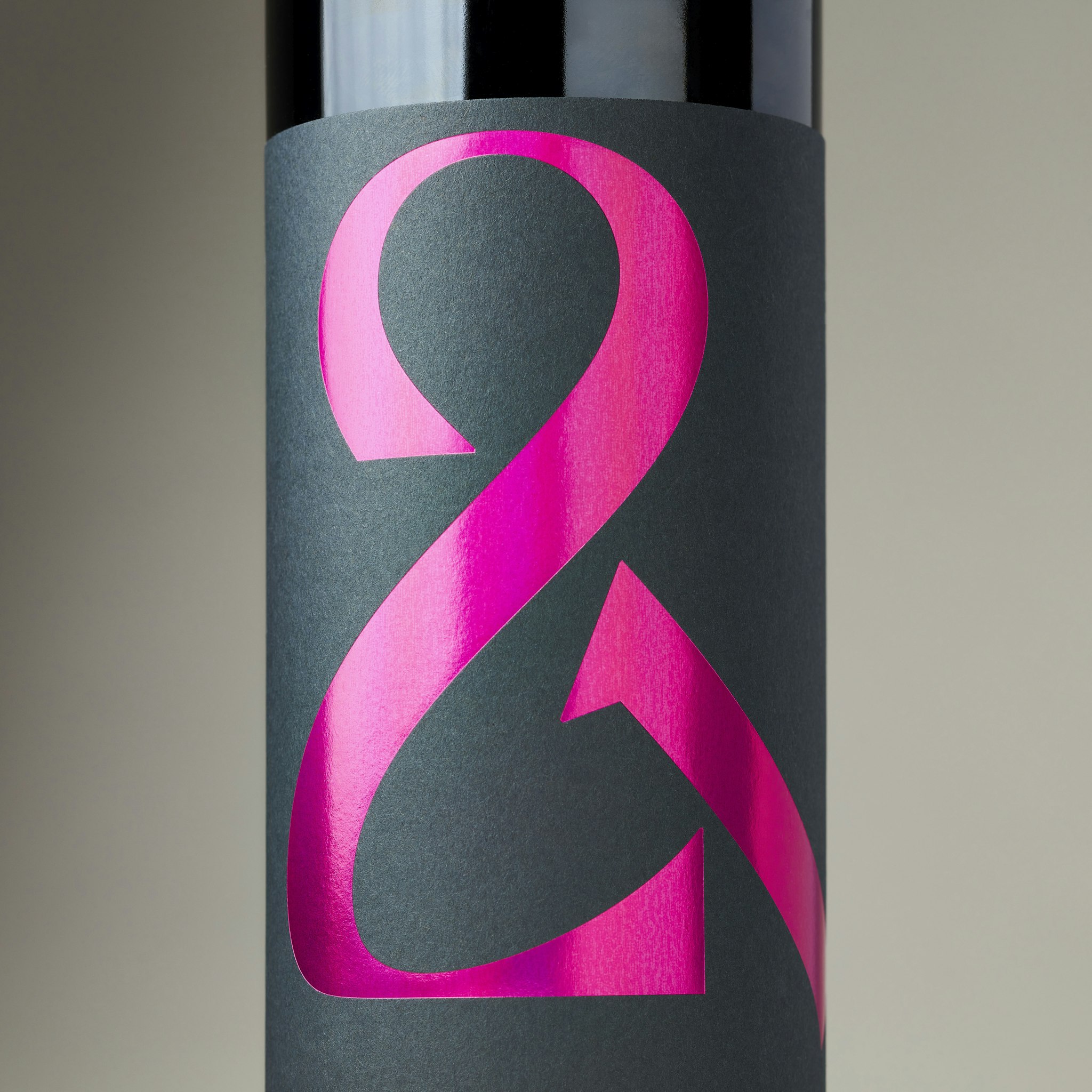 Close-up of red wine bottle label in dark grey with holographic hot pink ampersand logo under studio lighting