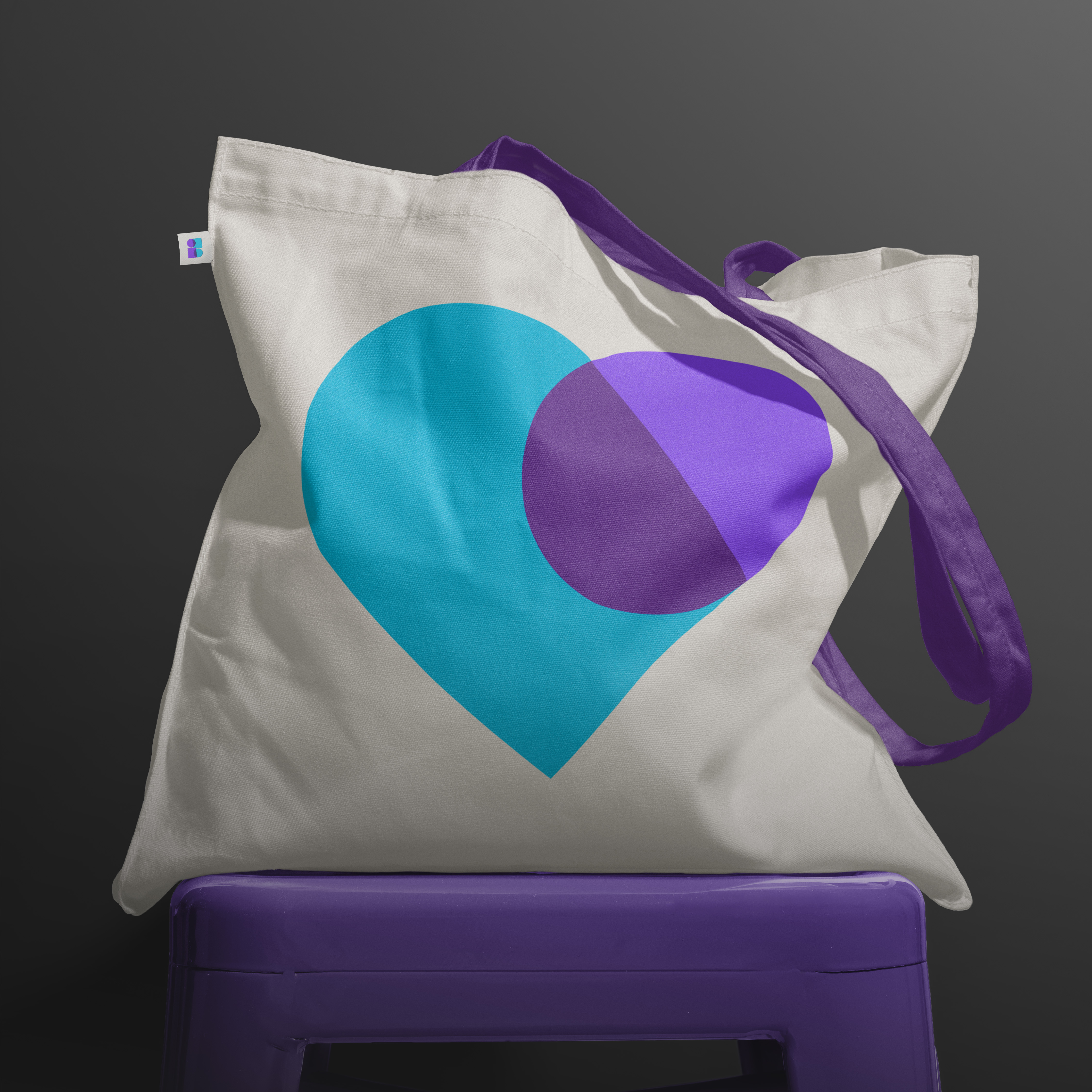 Cream tote bag with blue and purple abstract heart graphic and dark purple handles, filled and on dark purple stool against dark grey backdrop