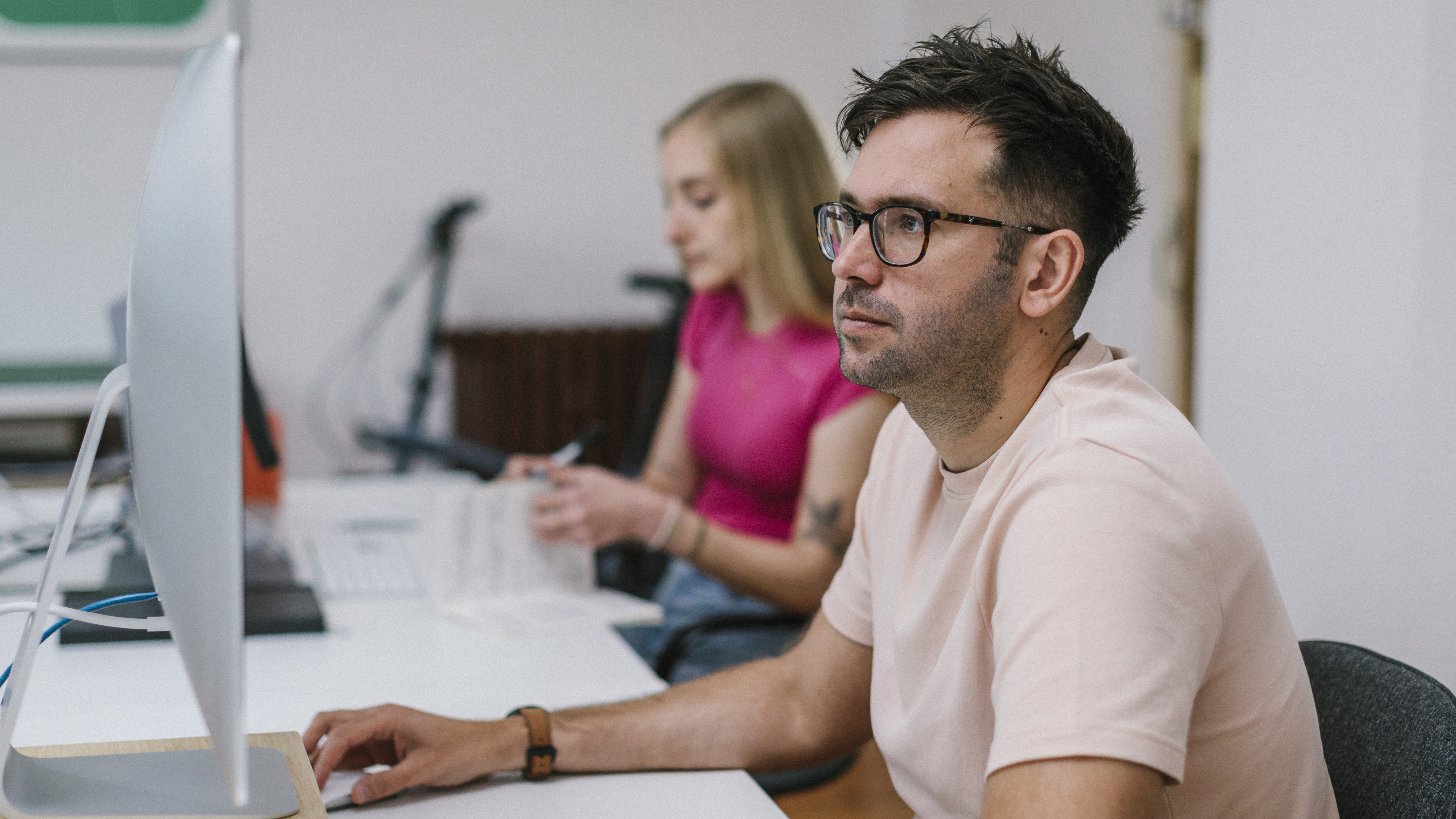 Focused man working on desktop computer in modern design studio with female colleague flicking through notebook in background