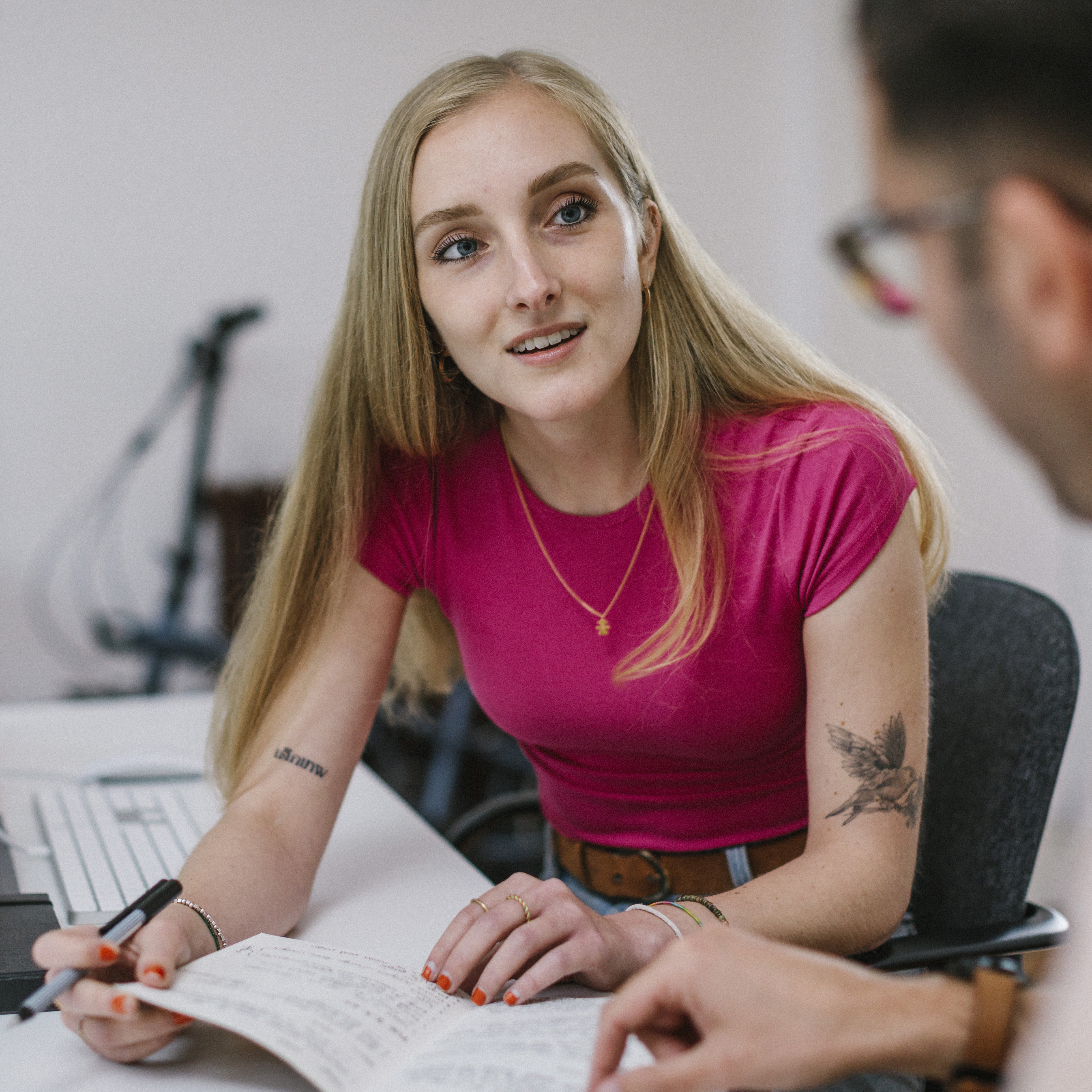 Young woman in pink t-shirt discussing ideas with colleague while taking notes in a modern design studio
