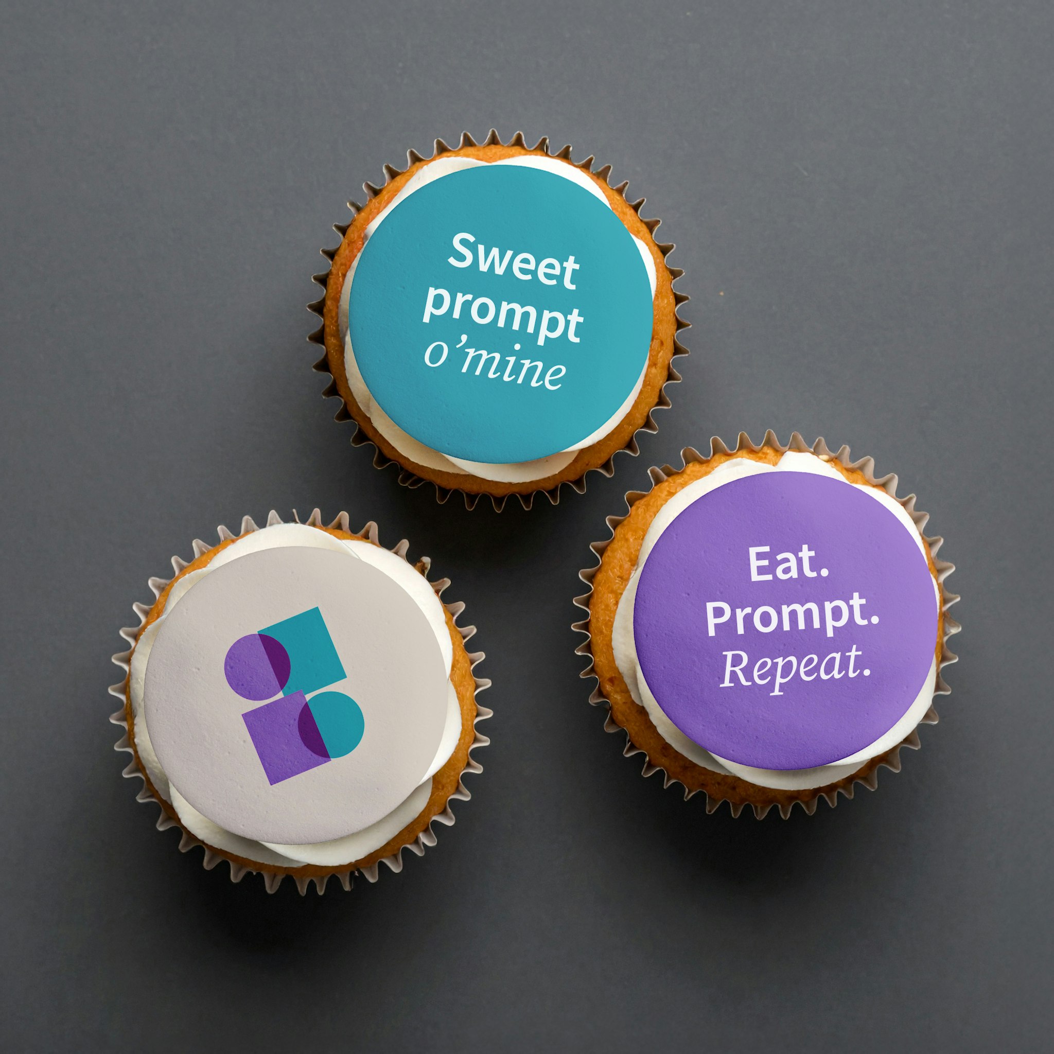 Three vanilla cupcakes on dark grey surface, one topped with IAG logo disk, two with purple and blue disks displaying campaign messages