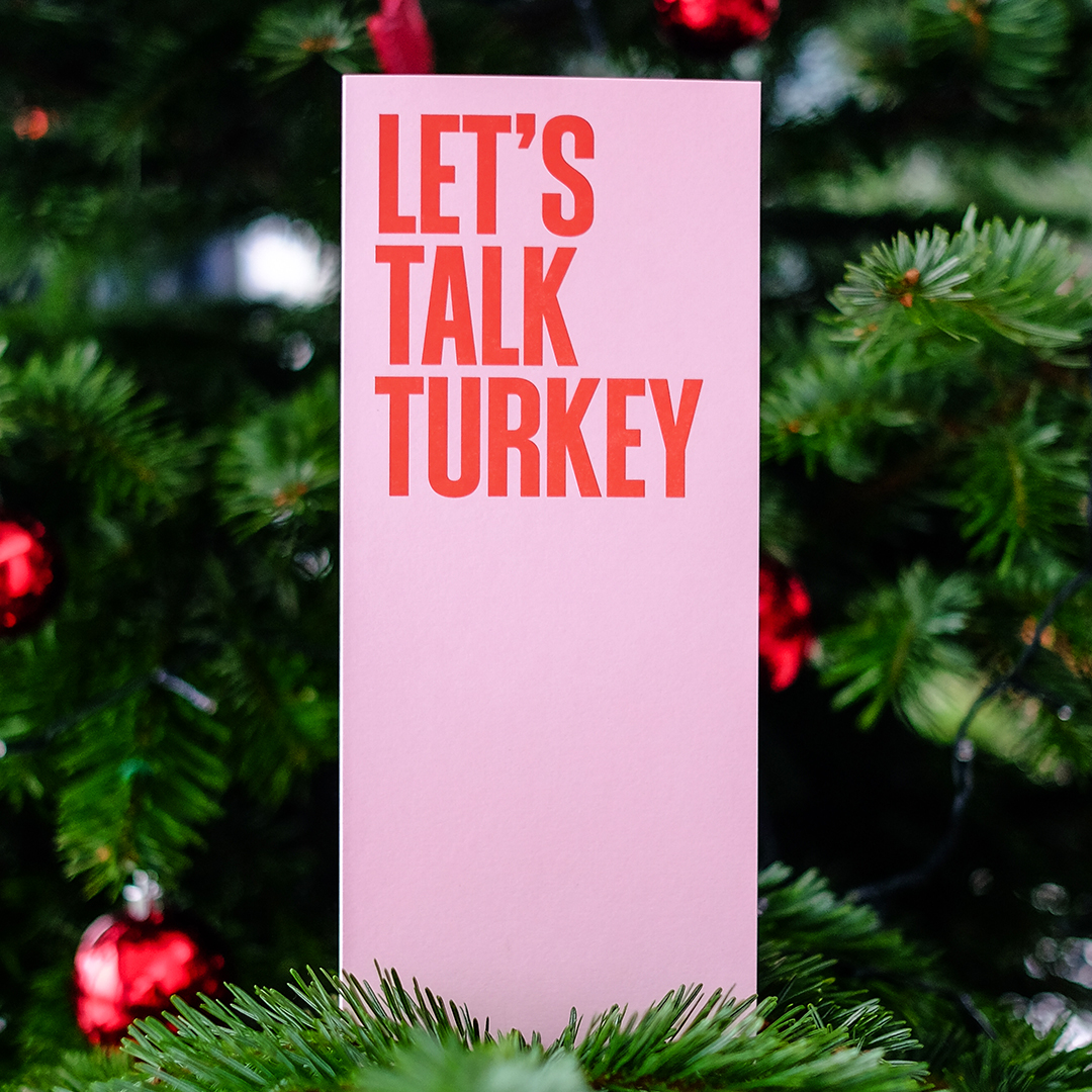 Pink Christmas card with bold red “Let’s Talk Turkey” on a branch in a decorated Christmas tree