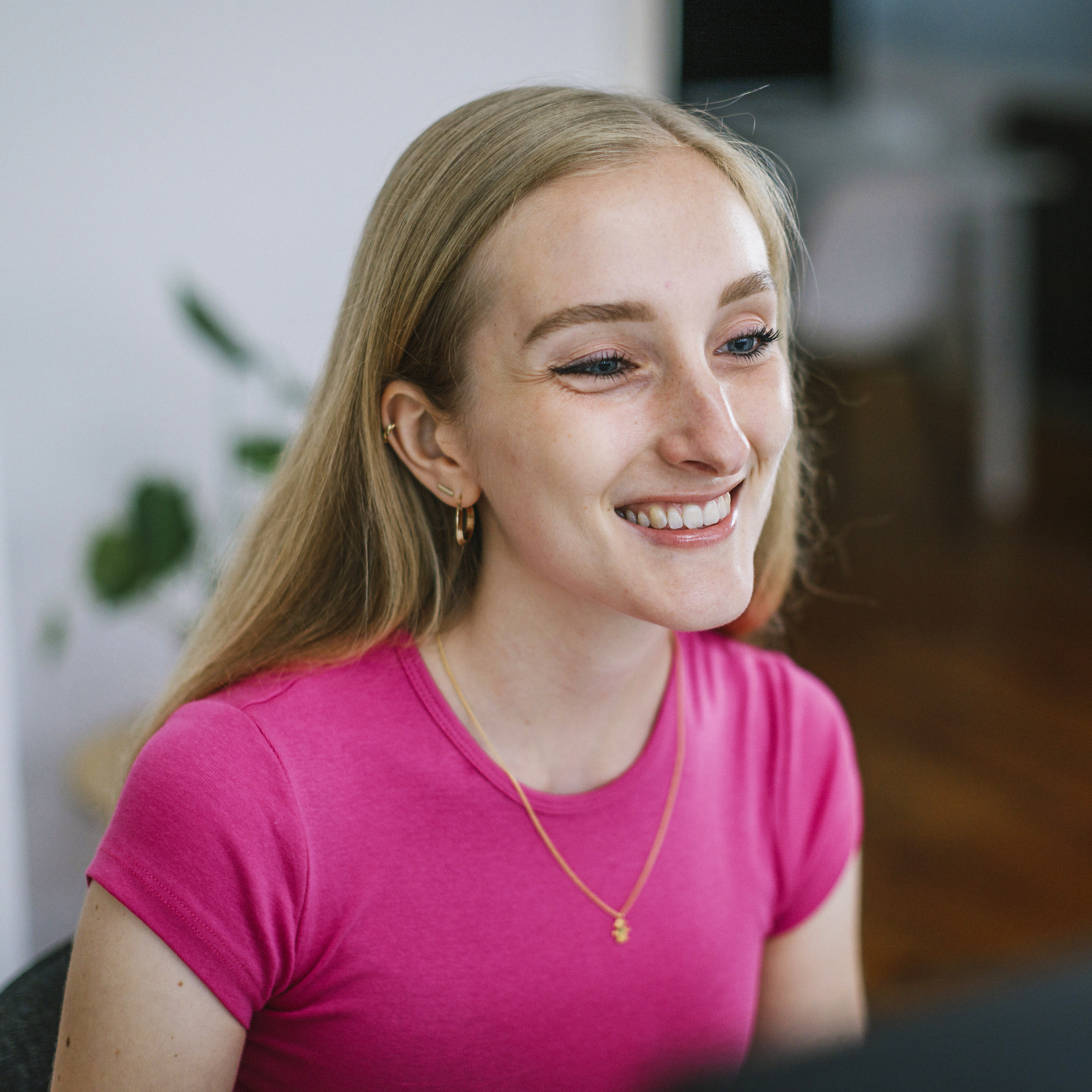 Young blonde woman in bright pink t-shirt sat at desk and smiling in colourful, modern design studio