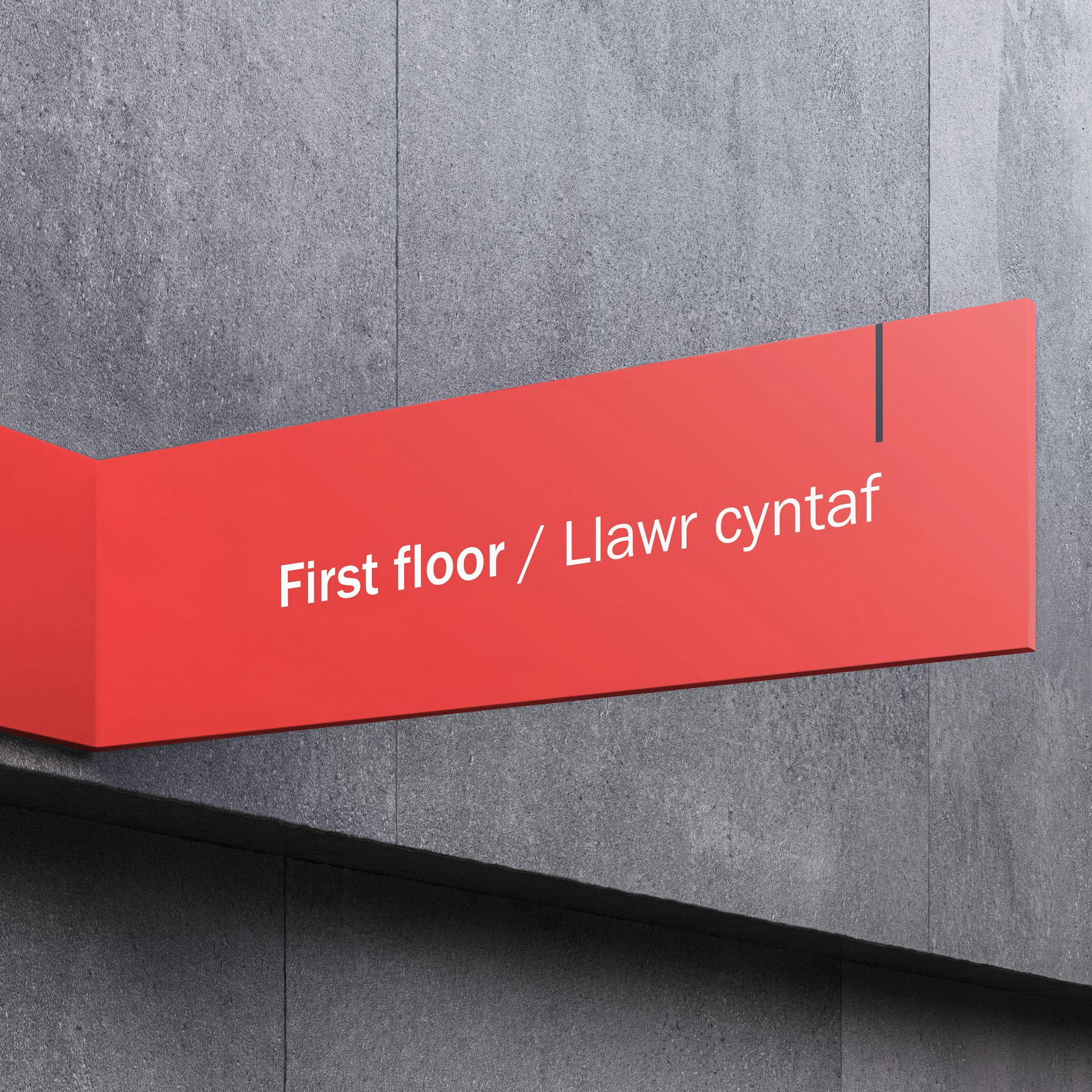 Red first floor sign with white text mounted on grey stone wall of modern building