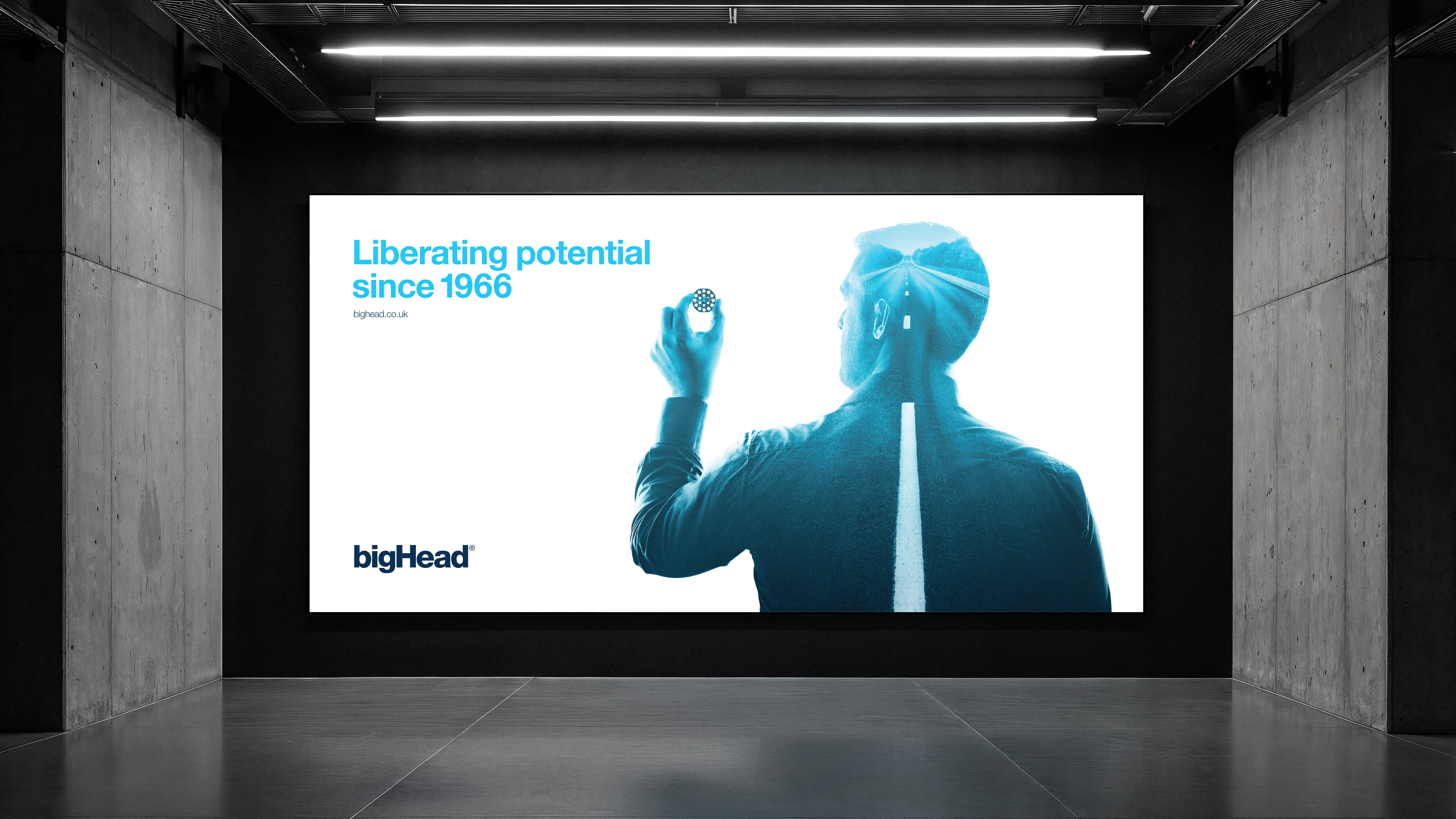 Digital billboard in industrial building showing blue double exposure of man holding bigHead with road image, logo and strapline in brand blues