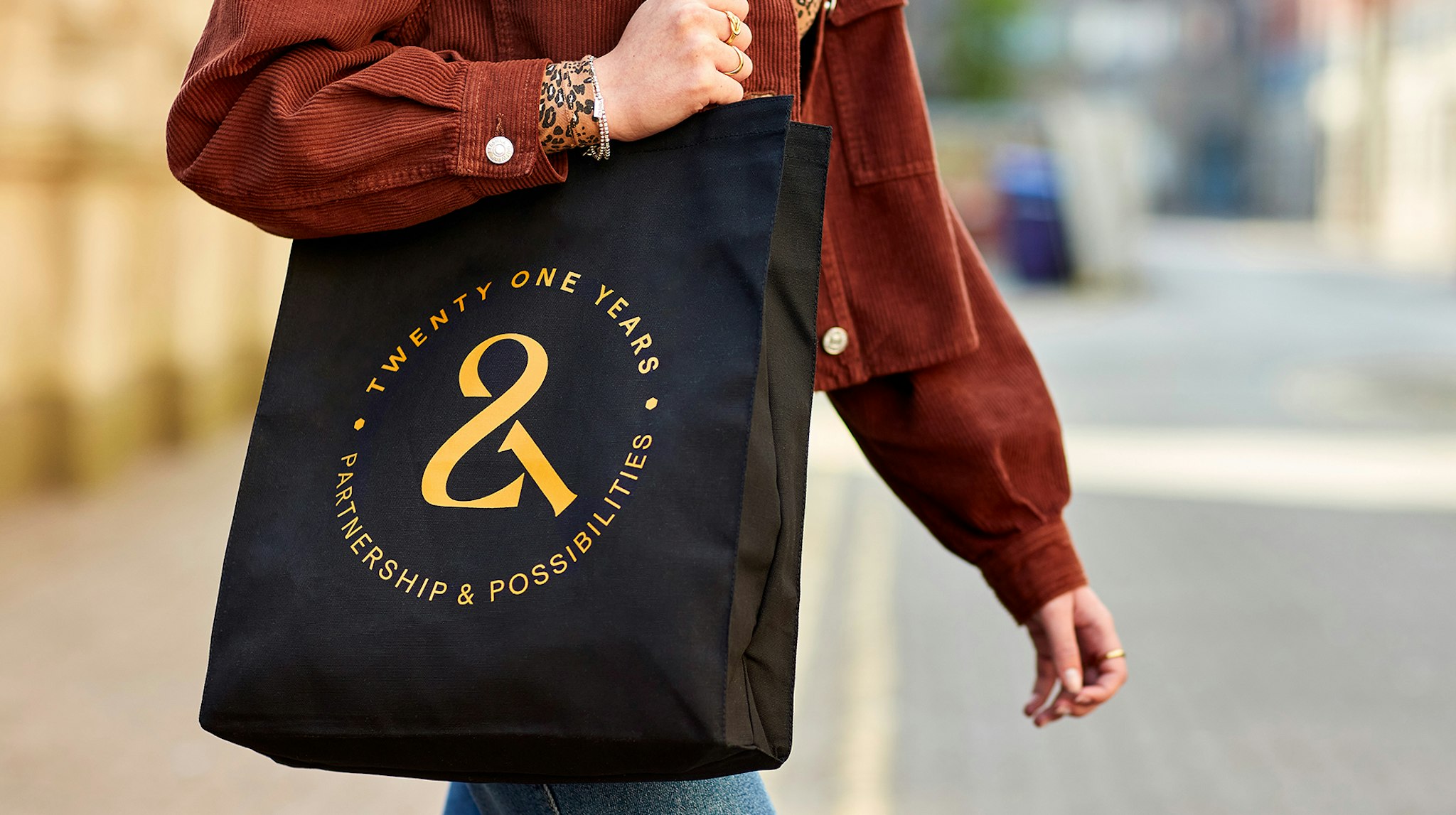 Person carrying a black tote bag with gold text reading 'Twenty One Years – Partnership & Possibilities' on a city street, celebrating a brand anniversary