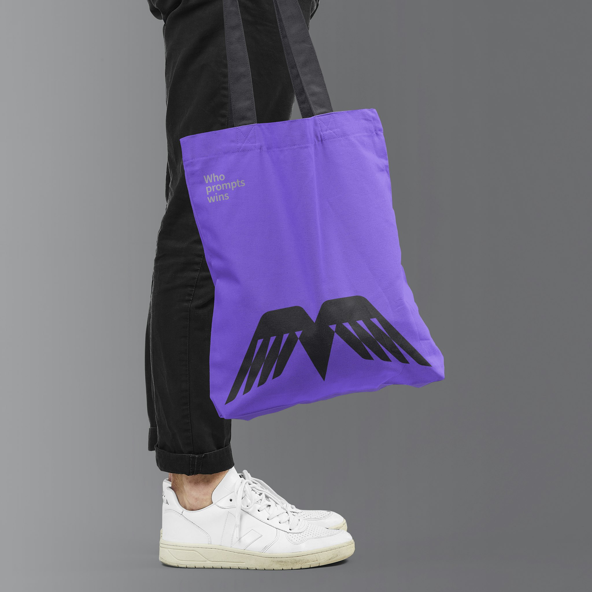 Man in black trousers and white trainers holding purple Prompt Mavericks tote with wing logo and silver campaign text against grey background