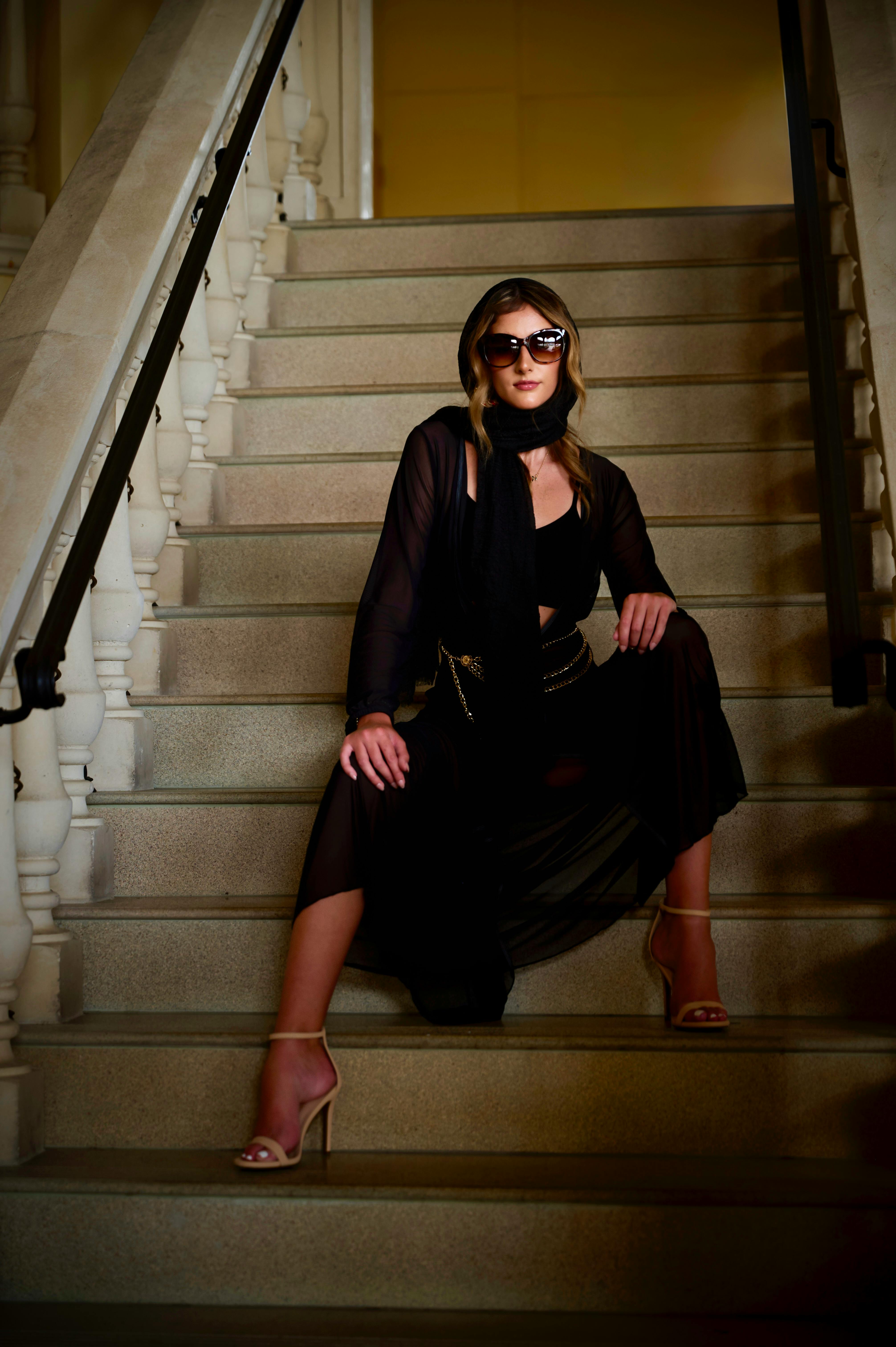Woman sitting on a stone staircase wearing a black dress and oversized sunglasses