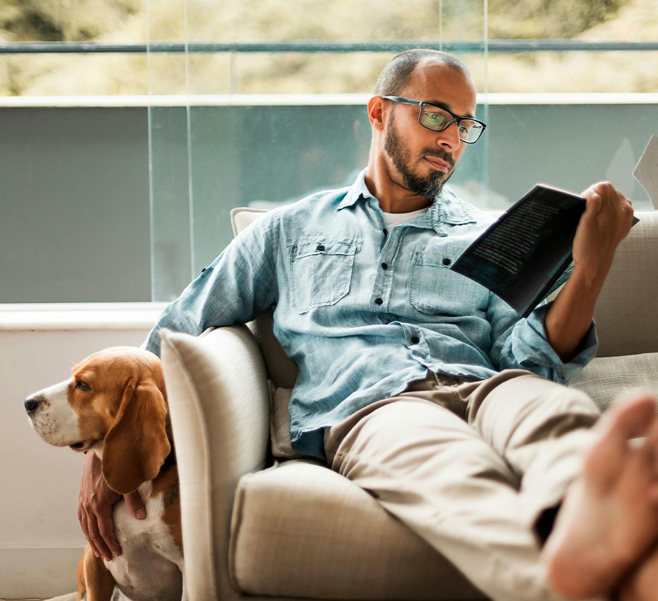 man reading book on couch with his feet kicked up while petting a dog