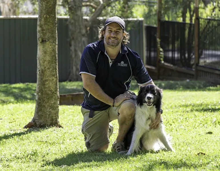 A Eumundi-Noosa dog boarding staff member sits with a border collie dog on the green grass
