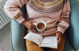 Foto en plano picado de una persona sentada en una silla, sosteniendo una taza de té y leyendo un libro.