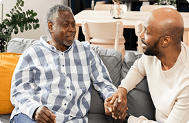 Dos hombres conversan en un sofá. El hombre más joven sostiene la mano del hombre mayor.