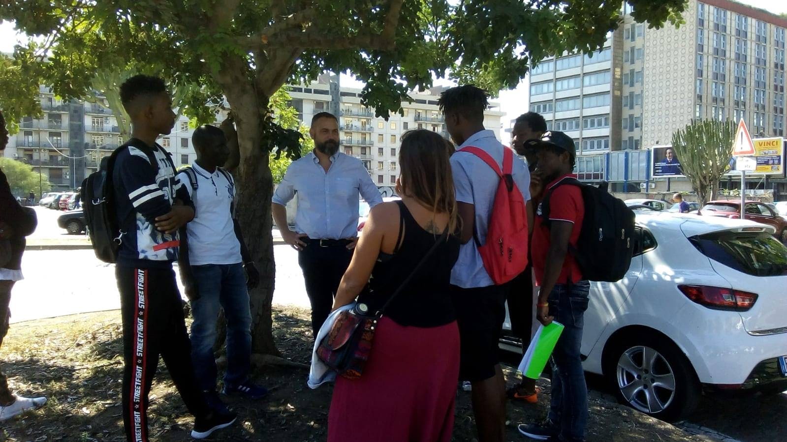 Under a carob tree, in an urban flowerbed, several people from different backgrounds share memories connected to the carob tree