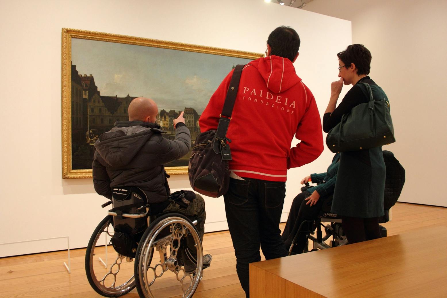 A group of visitors observes a work in a museum: the presence of people in wheelchairs and a staff member highlights how cultural accessibility involves not only space, but also the quality of relationships, guidance, and communication