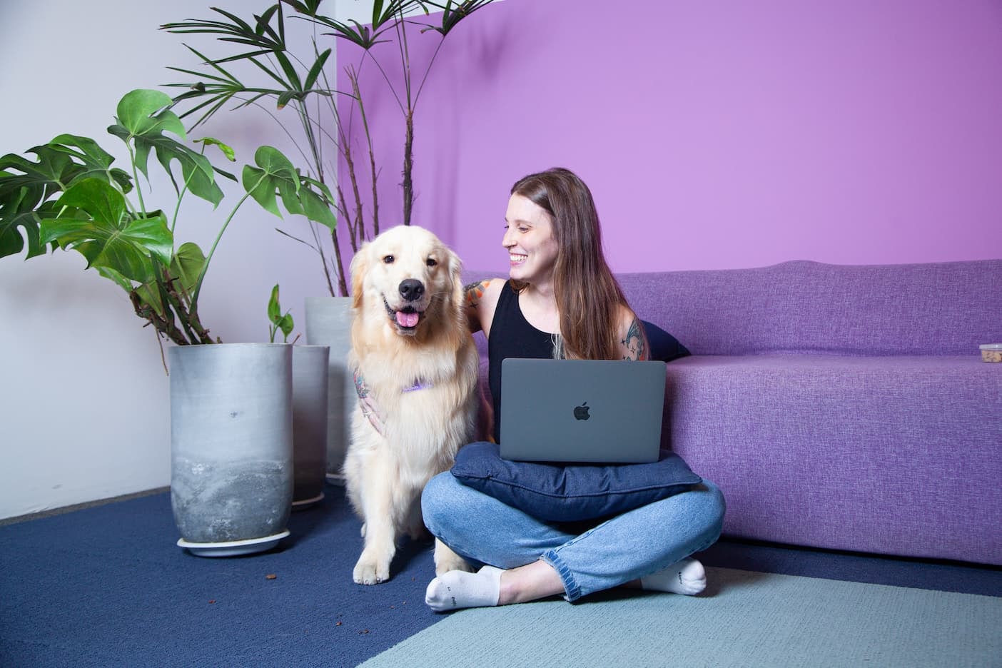 A woman sitting on the floor, leaning against a purple sofa, next to a seated Golden Retriever dog.