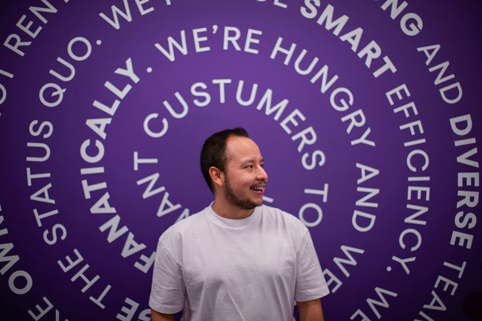 A smiling man in a white T-shirt in front of a purple wall with text arranged in a circular pattern.