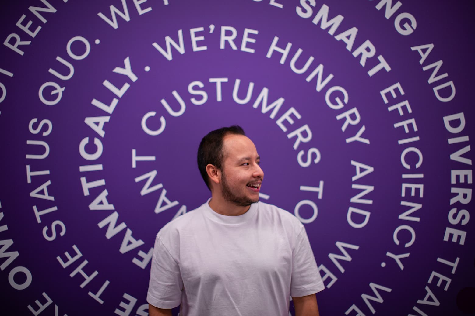 Hombre sonriente con camiseta blanca delante de una pared morada con frases en forma de círculo.