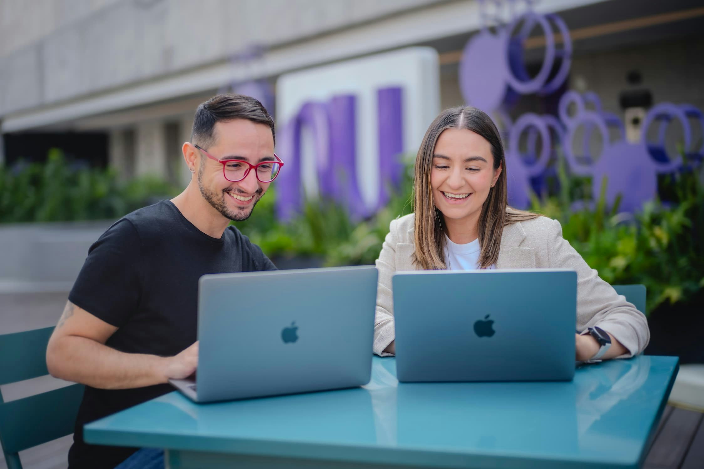 A man and a woman sitting at an outdoor table, using an Apple computer, with the Nu logo in the background.