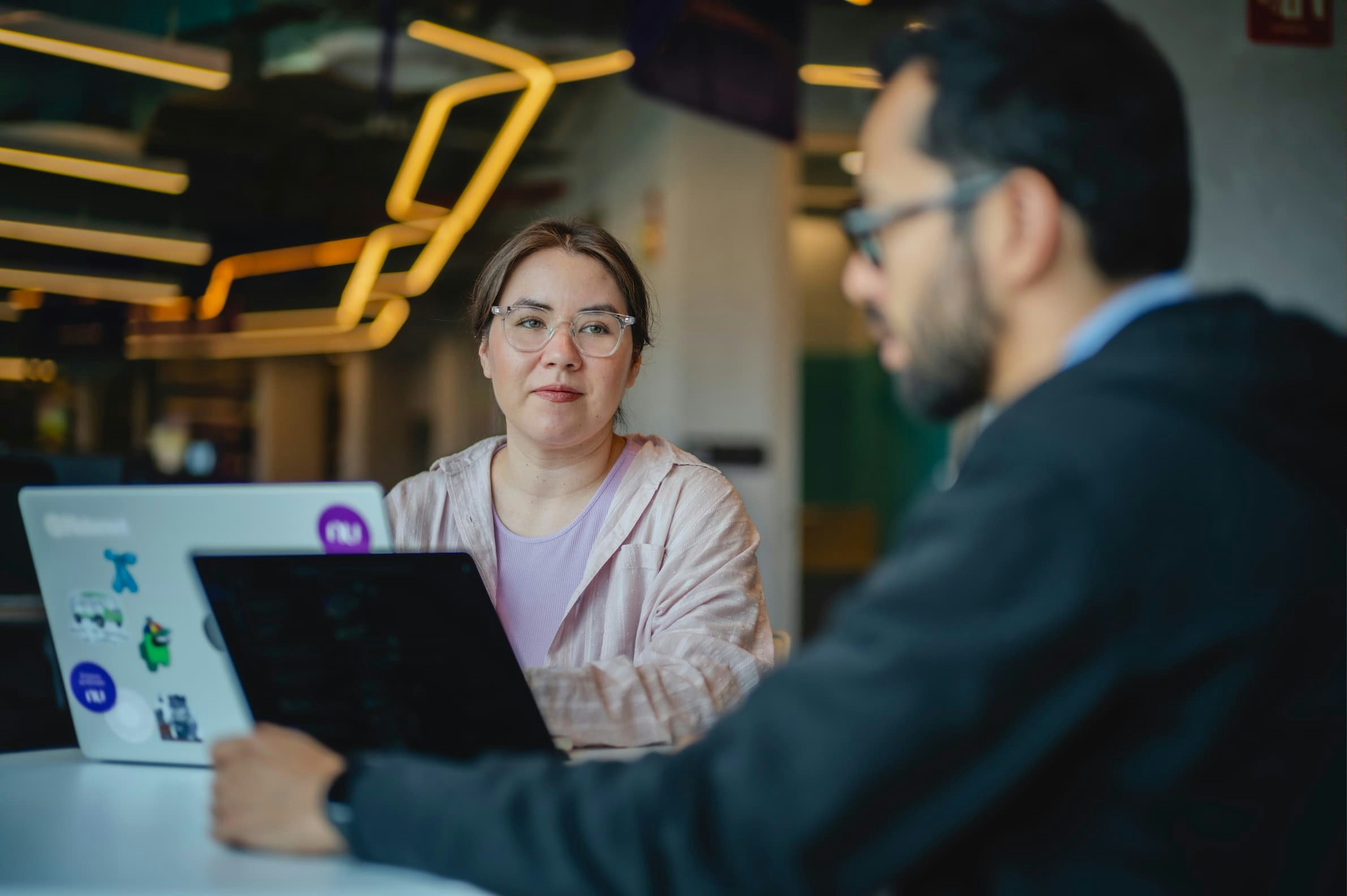 A woman wearing a lavender T-shirt and a bearded man sitting at a table in an office.