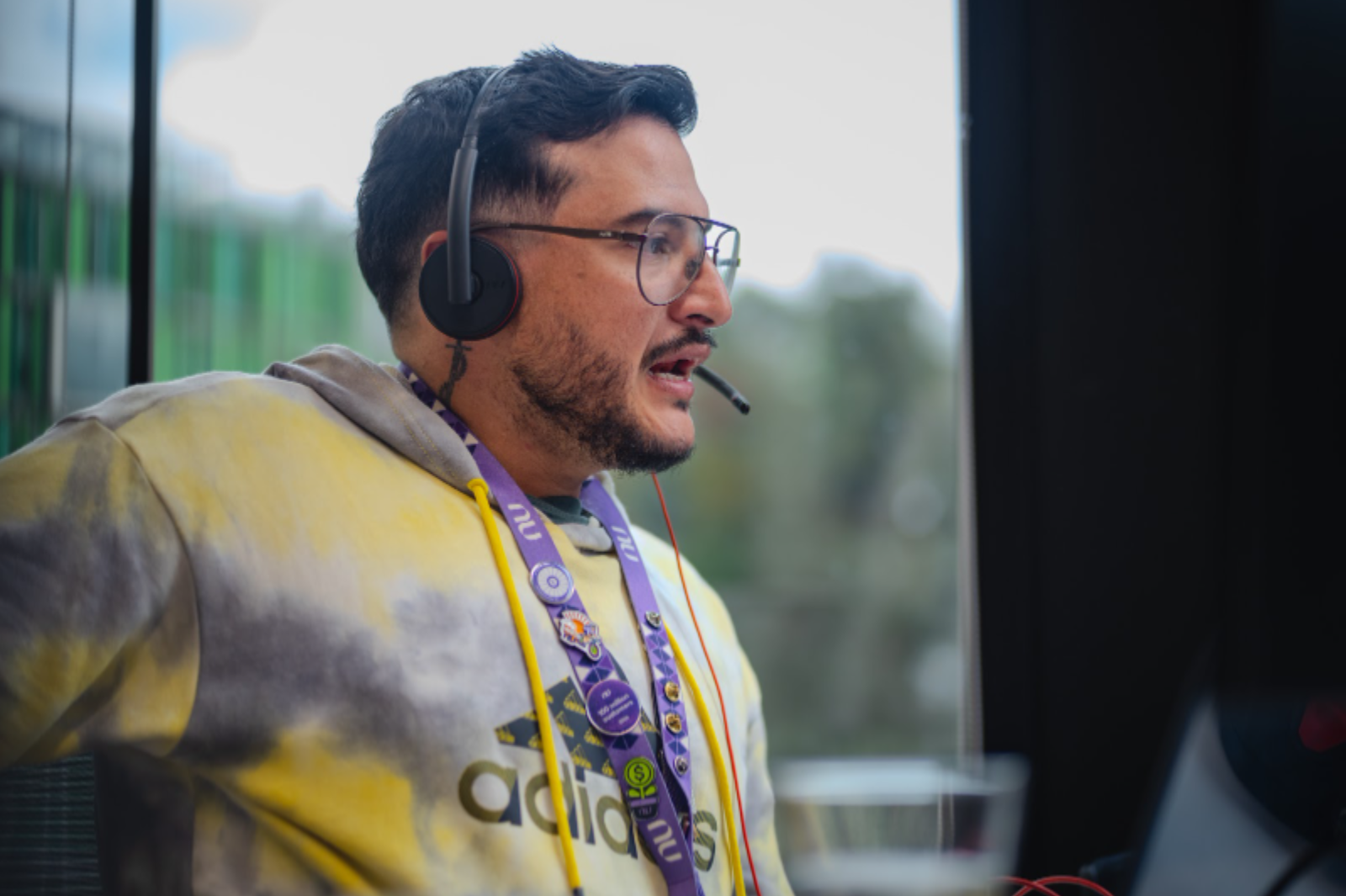 Hombre con gafas y auriculares, con cordón de Nubank, hablando frente a la computadora en la oficina.