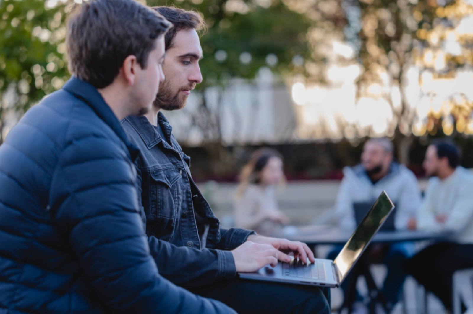 Two men sitting in an outdoor area looking at a laptop, with other people in the background.