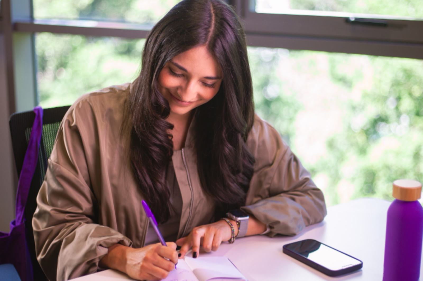 Woman smiling while writing in a notebook on a white table with a purple bottle, near a window.
