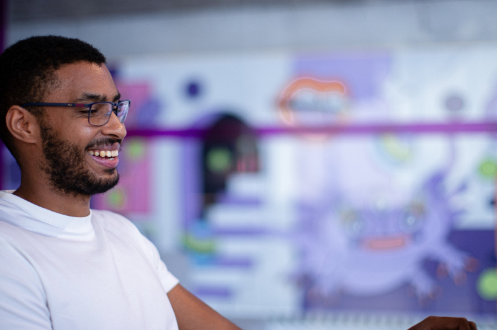 Hombre con gafas y camiseta blanca sonriendo mientras usa el portátil en una oficina con pared colorida.