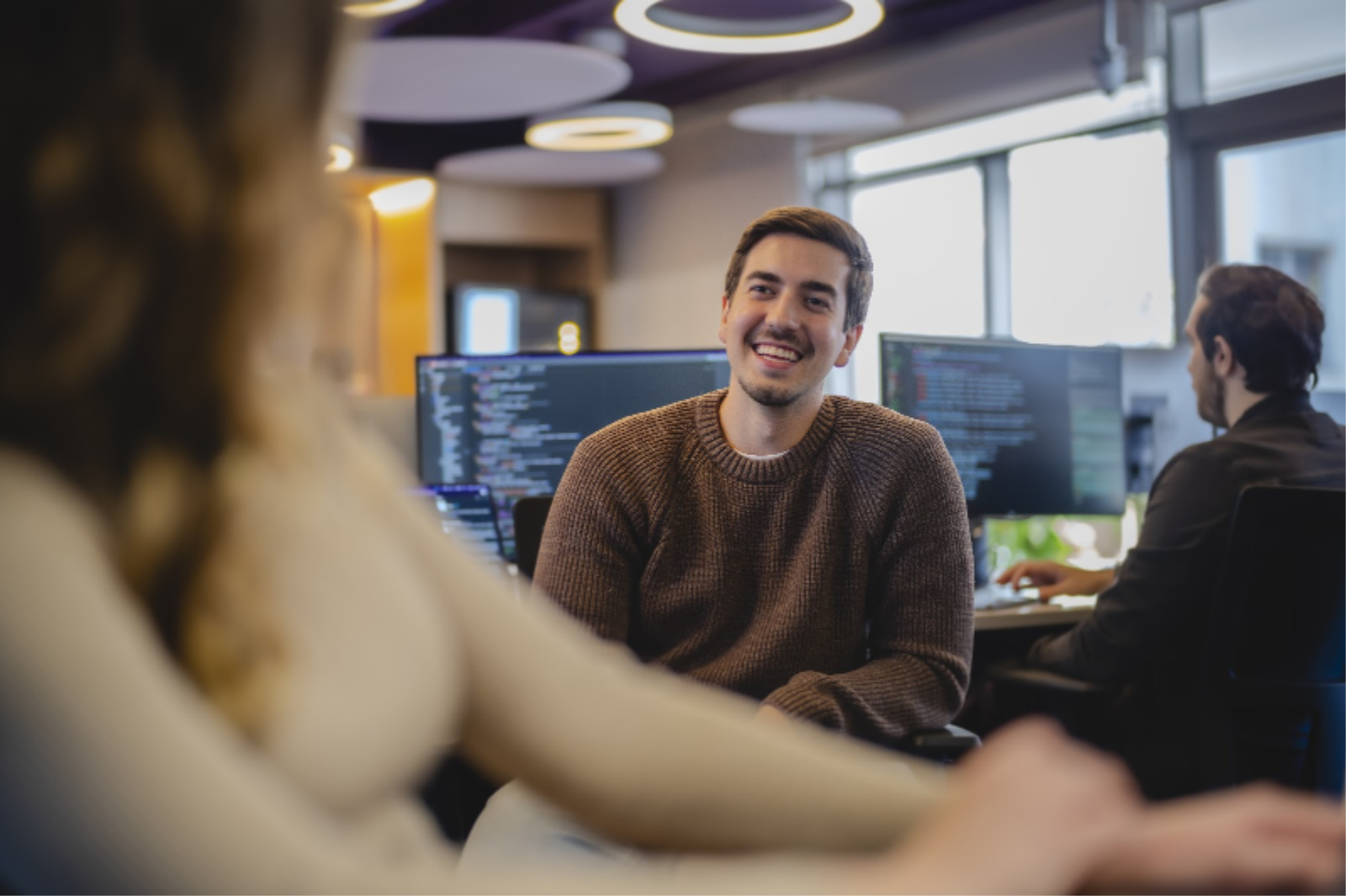 Hombre sonriendo conversando con un colega de trabajo en una oficina de tecnología con pantallas de código al fondo.