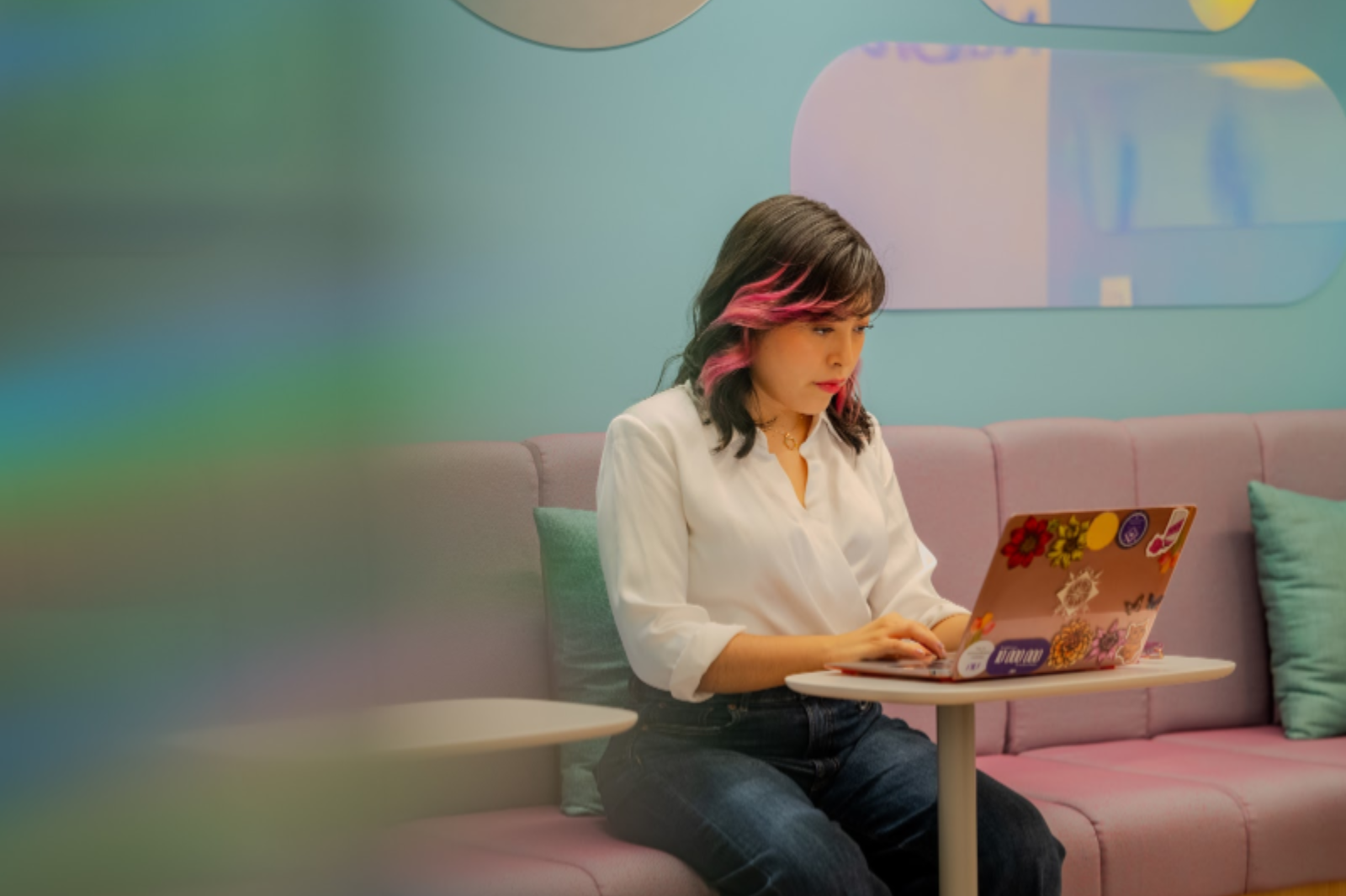 Mujer con mechones rosas en el pelo escribiendo en un portátil con pegatinas, sentada en un sofá lila.